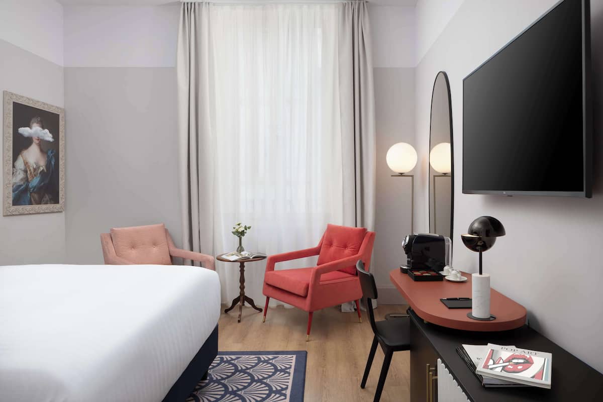 Ein Hotelzimmer mit einem Bett, einem roten Sessel, einem kleinen Tisch mit einer Vase, einem Schreibtisch mit einer Lampe und einem an der Wand hängenden Fernseher.