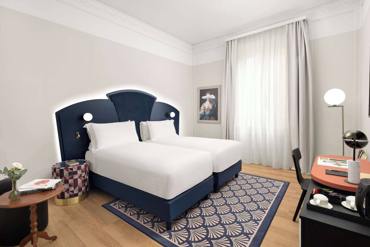 Ein Hotelzimmer mit einem Doppelbett, einem kleinen runden Tisch mit Lampe, einem Sessel, einem Nachttisch mit Vase und einem gemusterten Teppich.