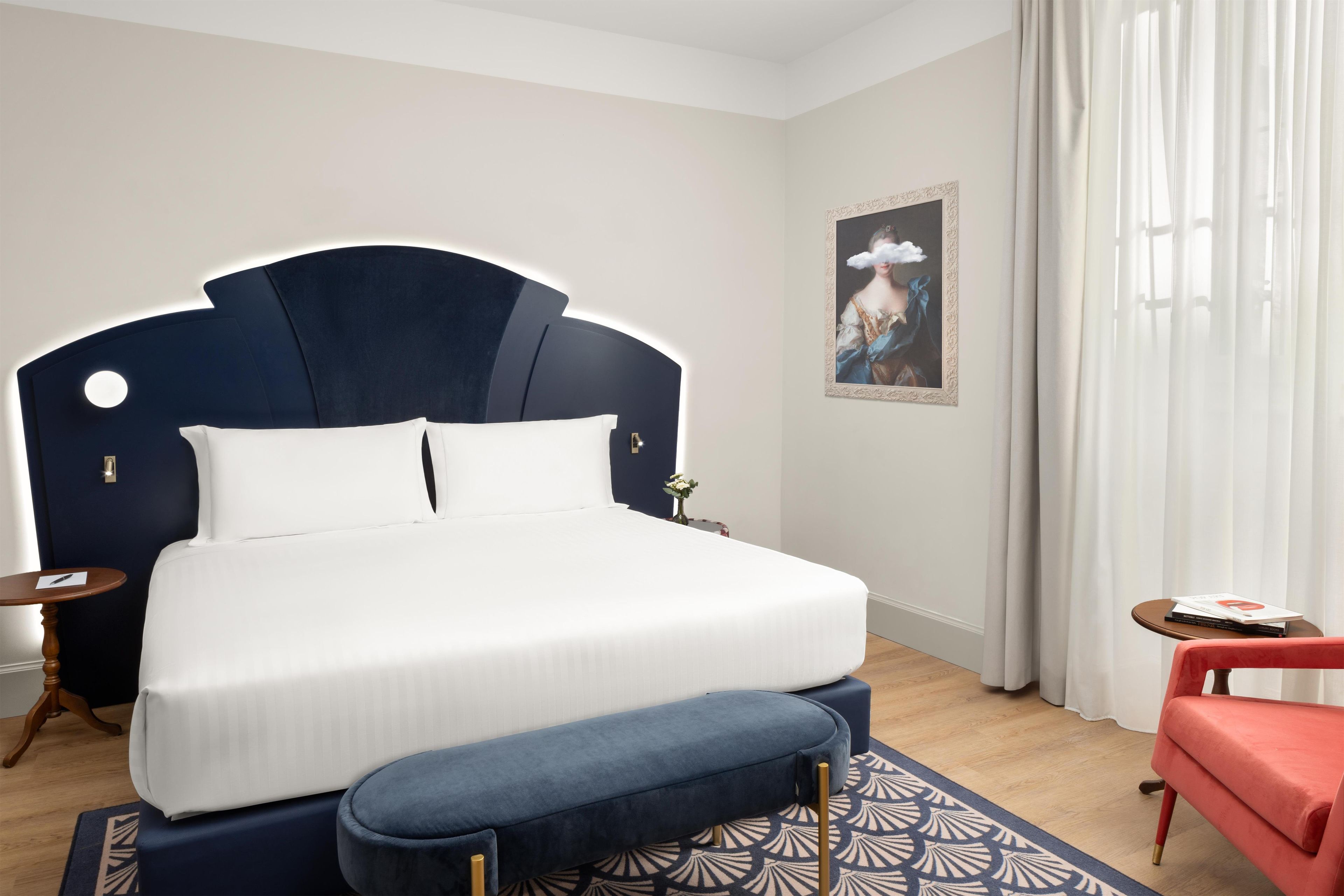 Une chambre d’hôtel bien rangée, avec un grand lit, une table de chevet, une petite table avec des livres, une chaise rouge et un tableau accroché au mur.
