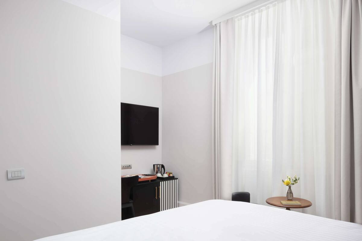Ein Hotelzimmer mit einem Bett, einem Schreibtisch mit Stuhl, einem an der Wand befestigten Fernseher und einem Fenster mit Vorhängen.