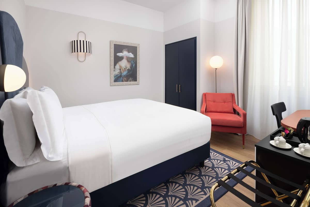Ein Hotelzimmer mit einem Bett, einem roten Sessel, einem Schreibtisch mit einer Lampe und einem Gemälde an der Wand.