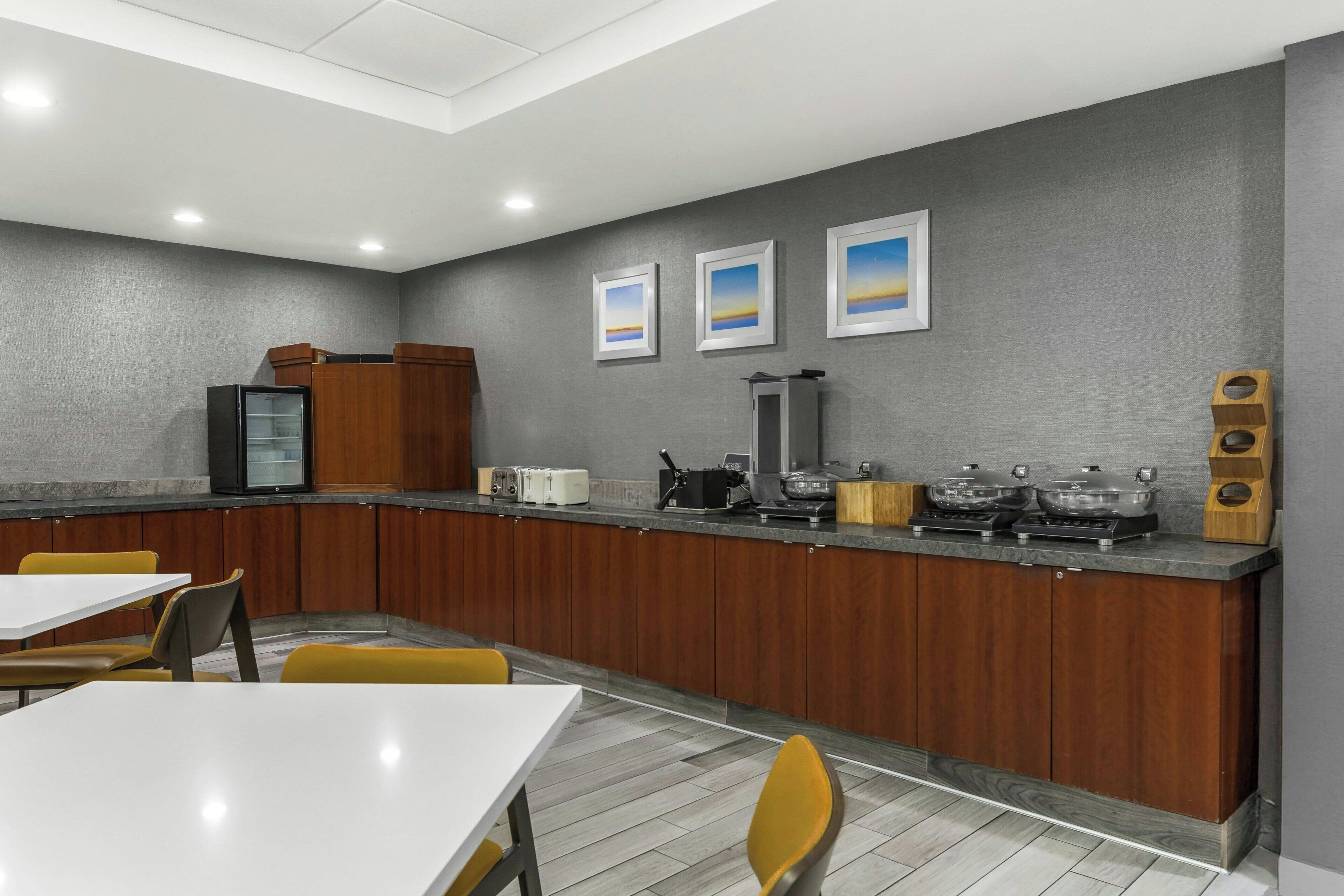 A modern kitchen with a grey wall, wooden cabinets, and a counter with appliances. There are yellow chairs and a white table.