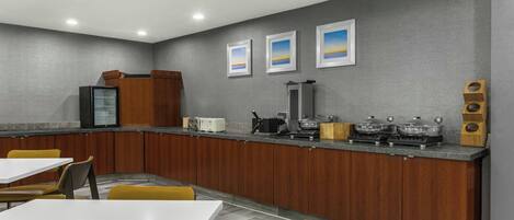 A modern kitchen with a grey wall, wooden cabinets, and a counter with appliances. There are yellow chairs and a white table.