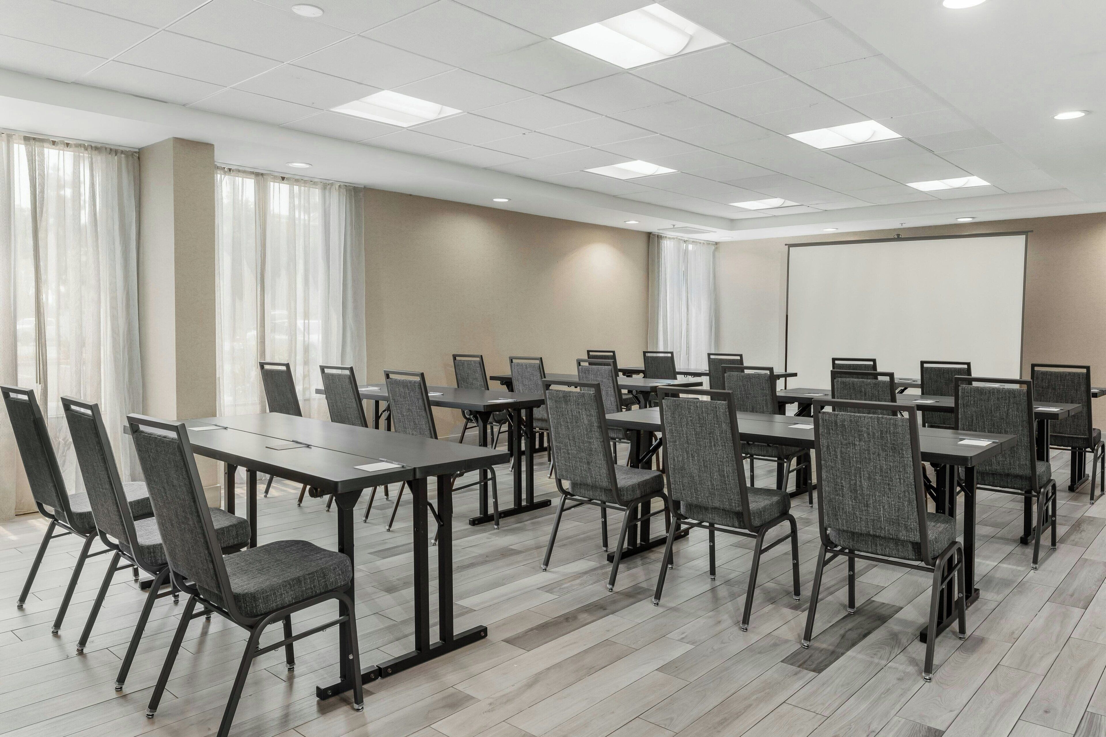 A conference room with multiple tables and chairs, a projector screen, and a large window with curtains.
