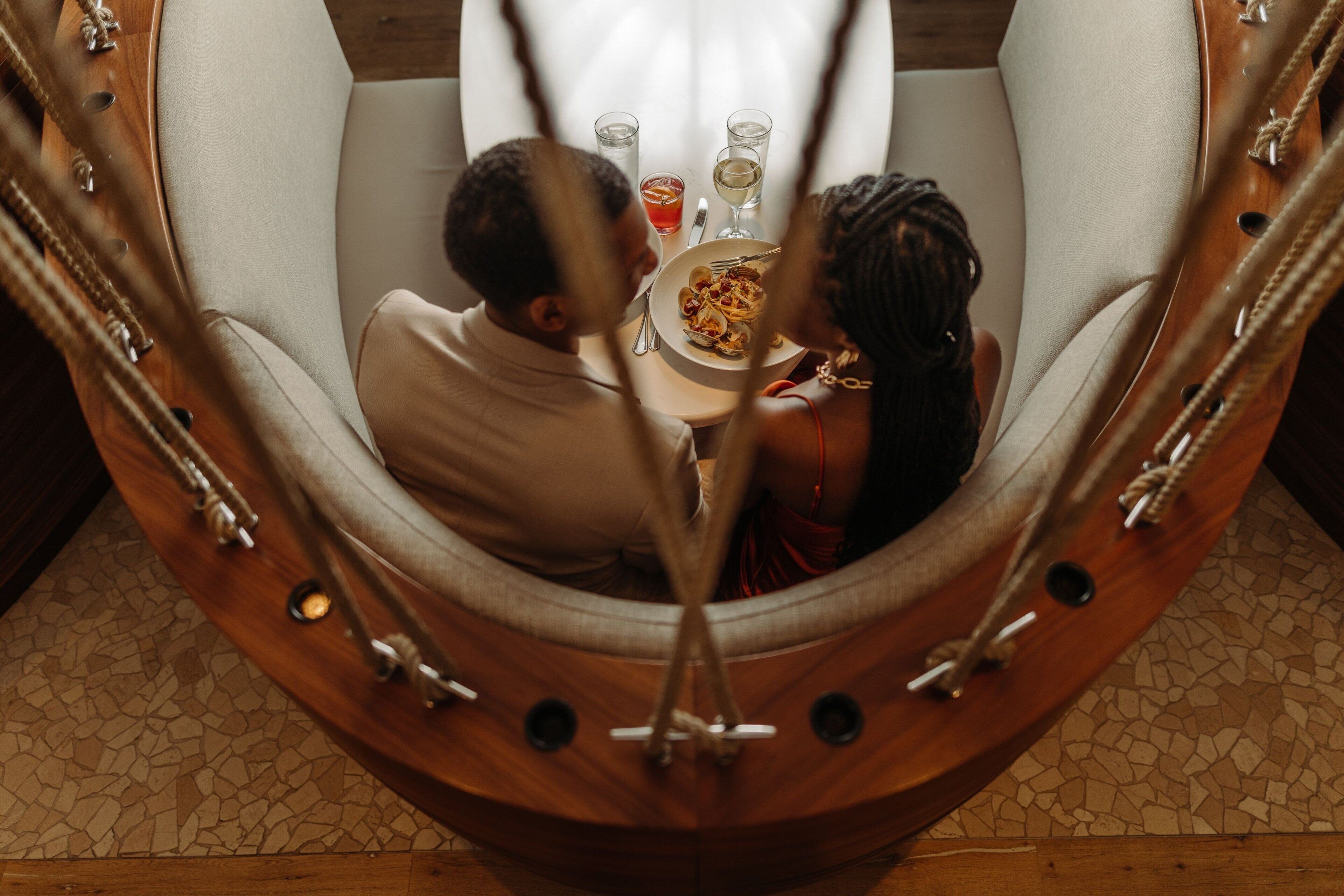 A couple dining in a hammock chair with a table set for two, a glass of wine, and a plate of food.