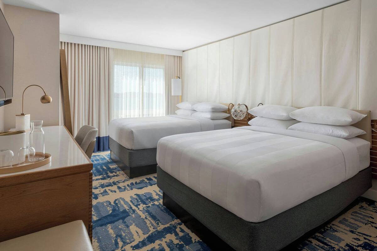 A hotel room with two beds, a desk, a chair, a lamp, and a window with curtains.