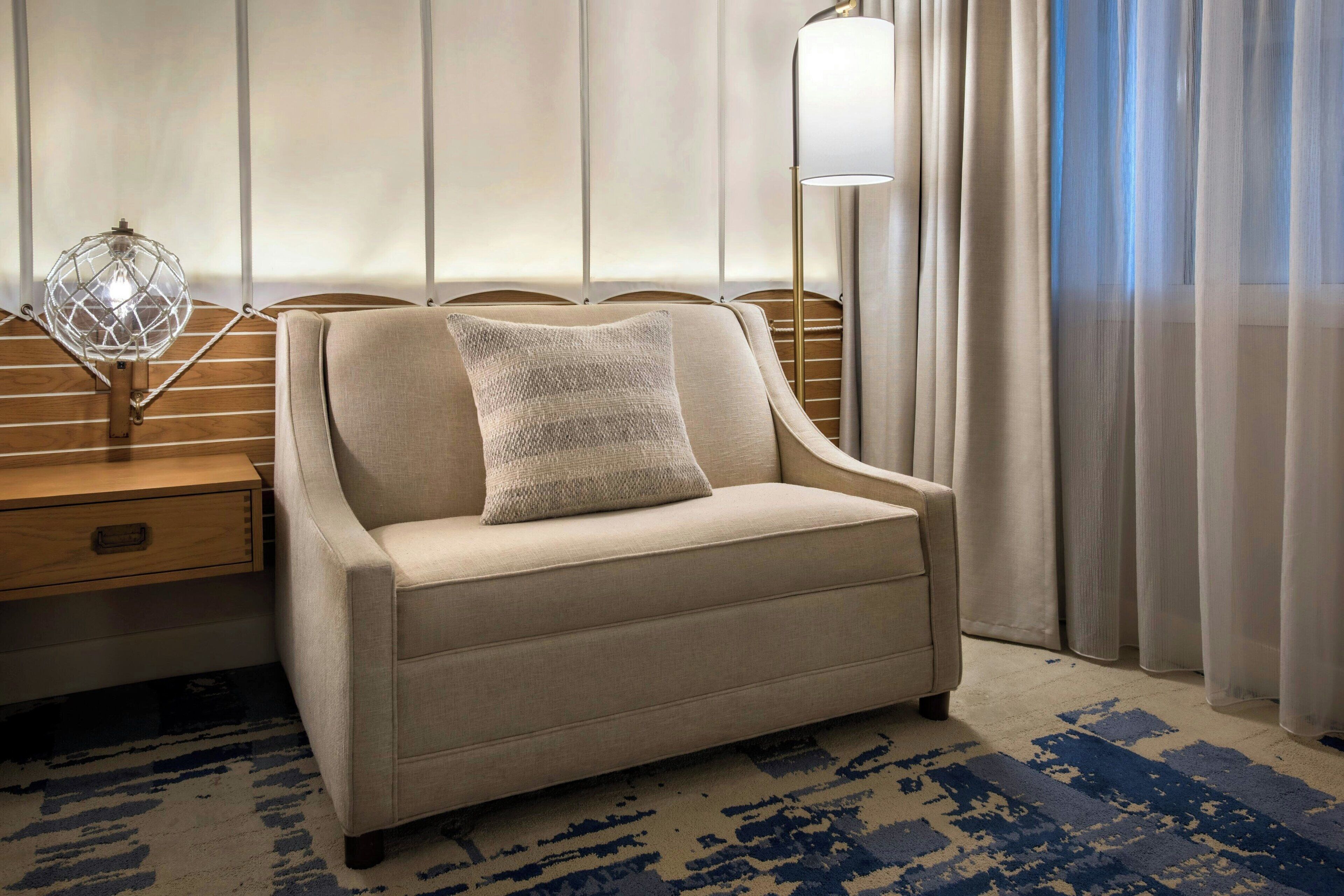A hotel room with a beige sofa, a decorative lamp, a wooden nightstand, and a patterned rug.