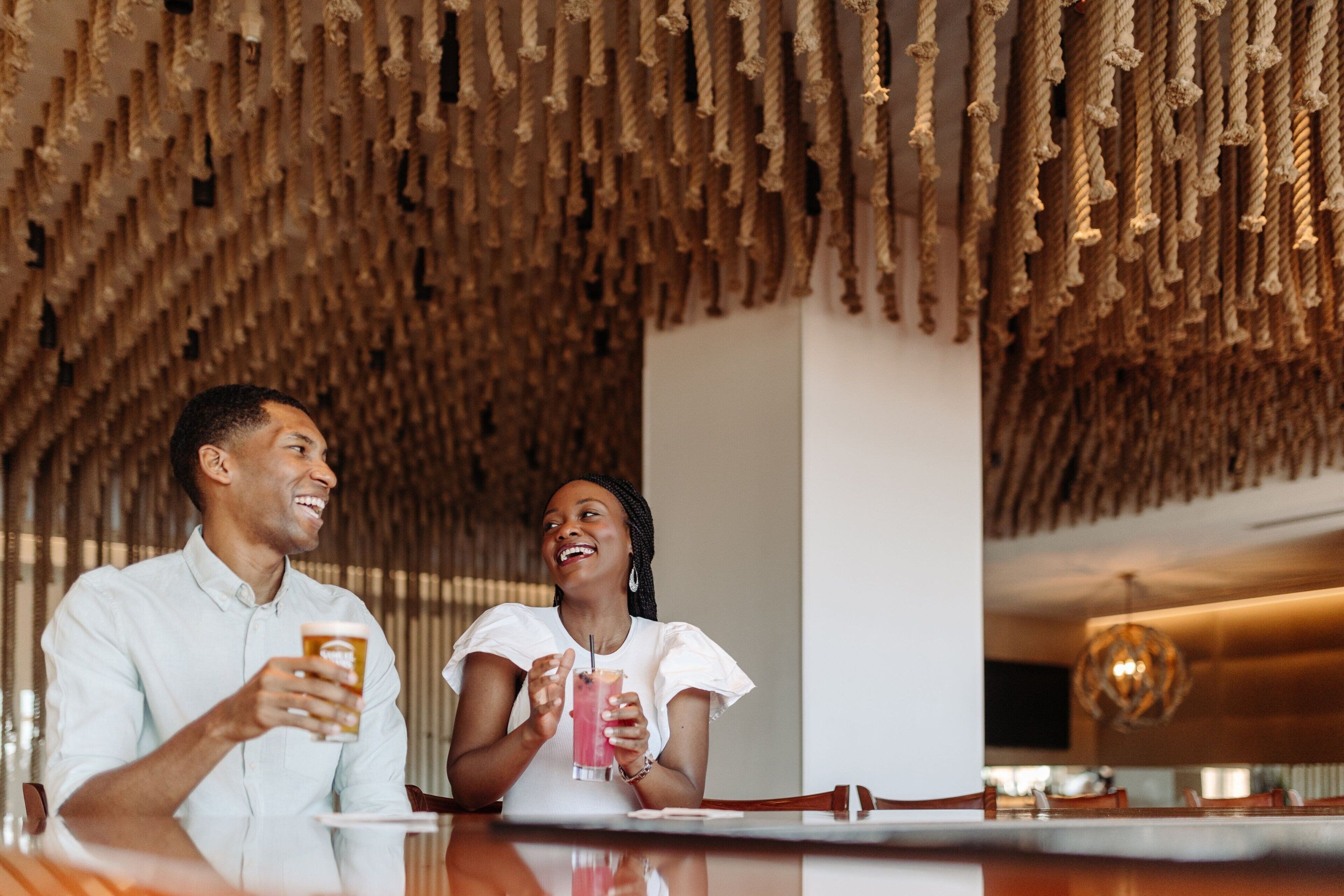 A couple sitting at a bar with a unique ceiling made of hanging ropes, a modern chandelier, and a clean, minimalist interior.