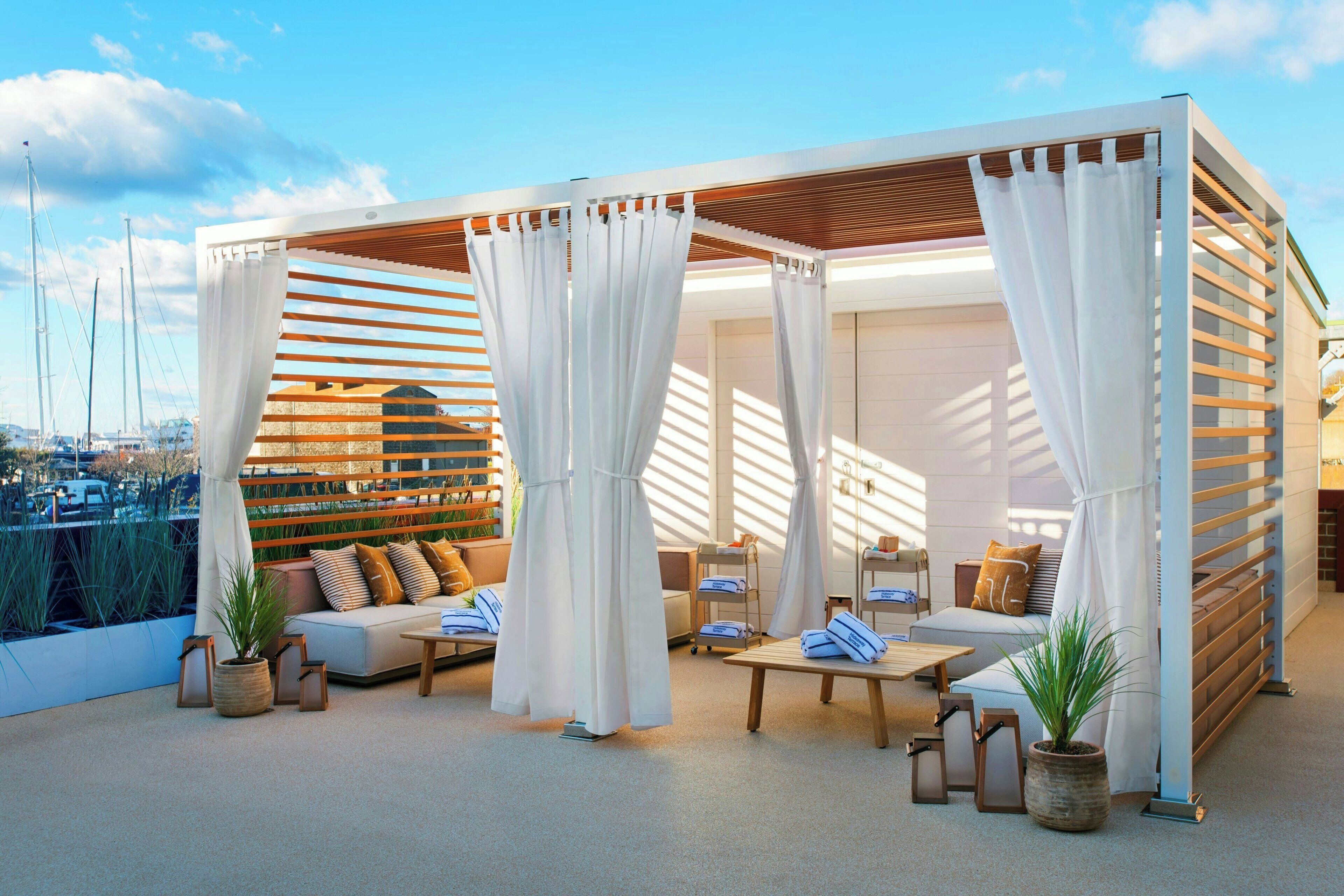 Un salón en la azotea con una pérgola de madera, cortinas blancas y un espacio de estar con cojines y una mesa de centro.
