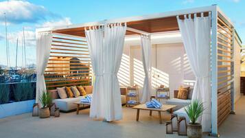 Un salón en la azotea con una pérgola de madera, cortinas blancas y un espacio de estar con cojines y una mesa de centro.