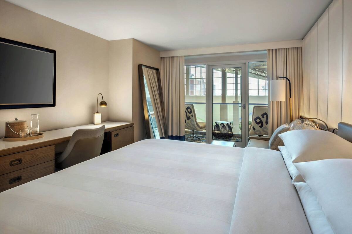 A hotel room with a large bed, a desk with a lamp, a chair, and a view of the outdoors through a large window.