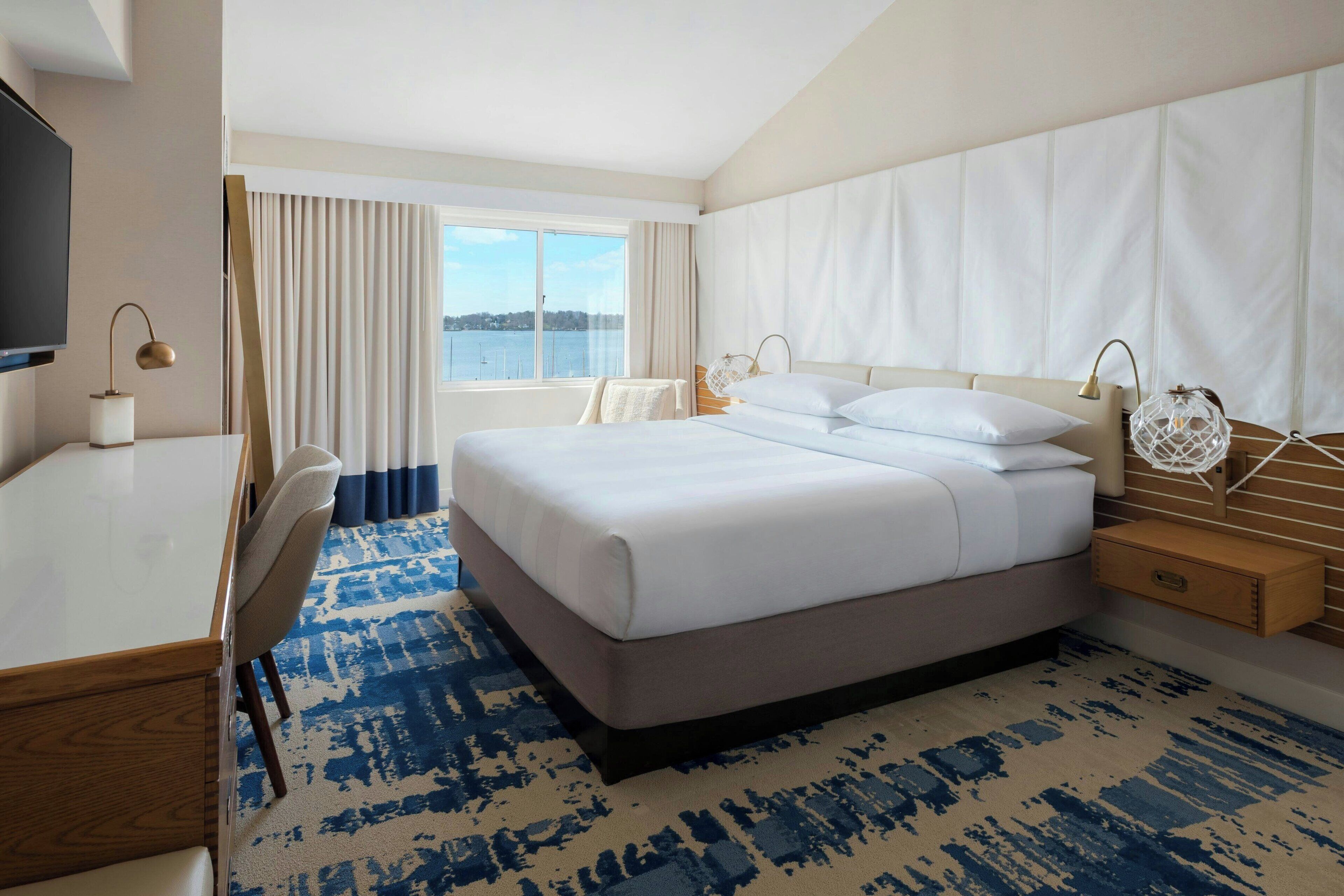 Quarto de hotel com uma cama grande, uma escrivaninha com cadeira, televisão e janela com cortinas.