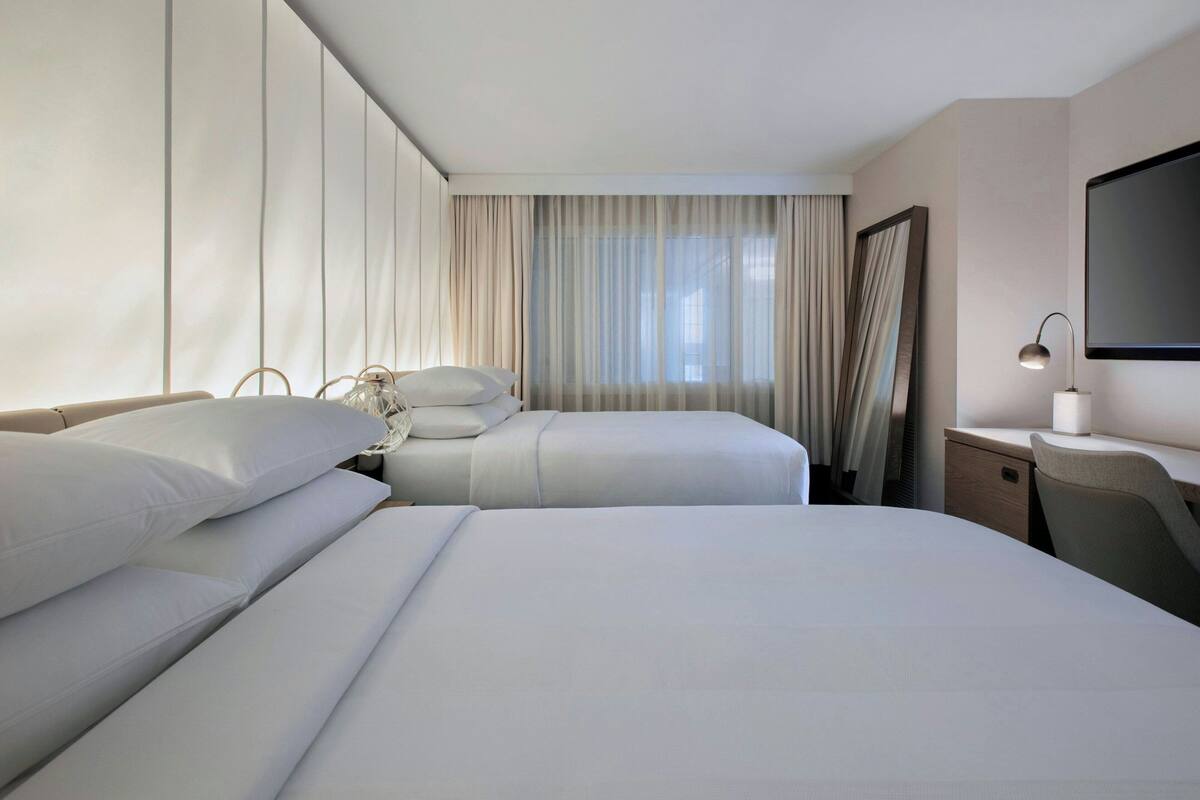 A hotel room with two beds, a desk, a chair, a TV, and a window with curtains.