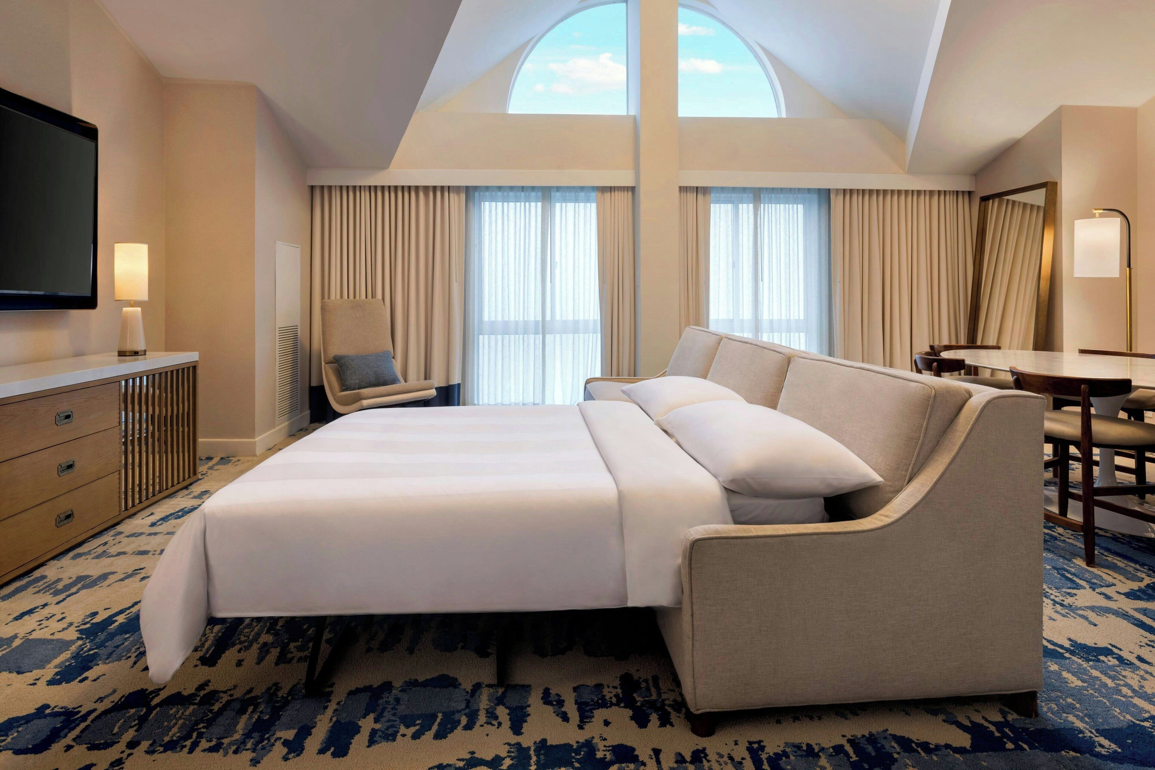 A hotel room with a large bed, a desk, a chair, a TV, and a large window with curtains.