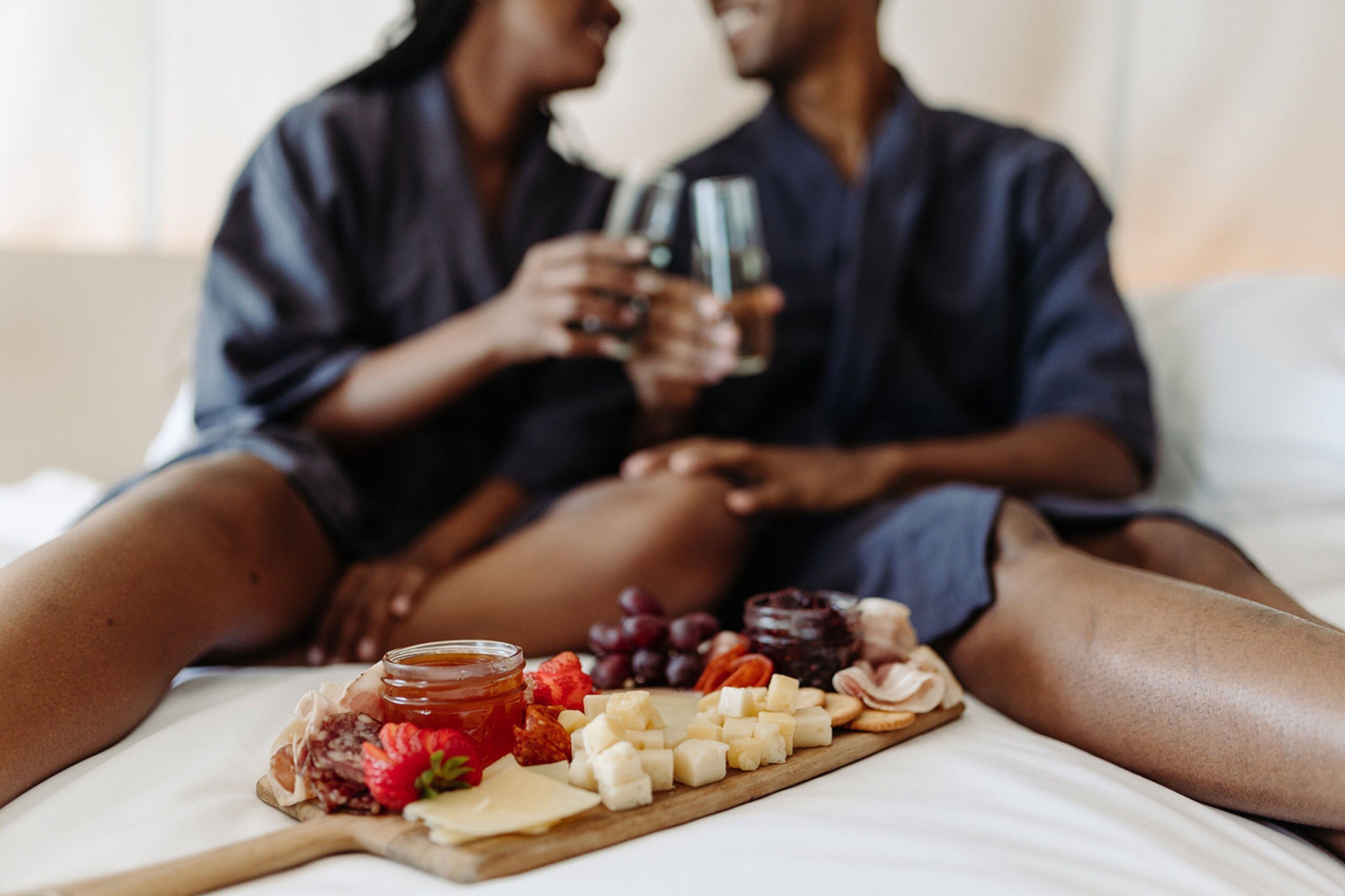 Una pareja sentada en una cama con una tabla de quesos delante.