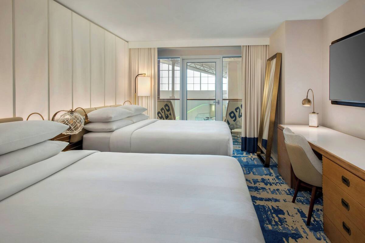 A hotel room with two beds, a desk, a TV, and a view of the outside.