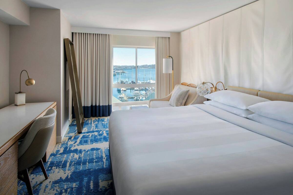 A hotel room with a large bed, a desk, a chair, and a view of the marina.