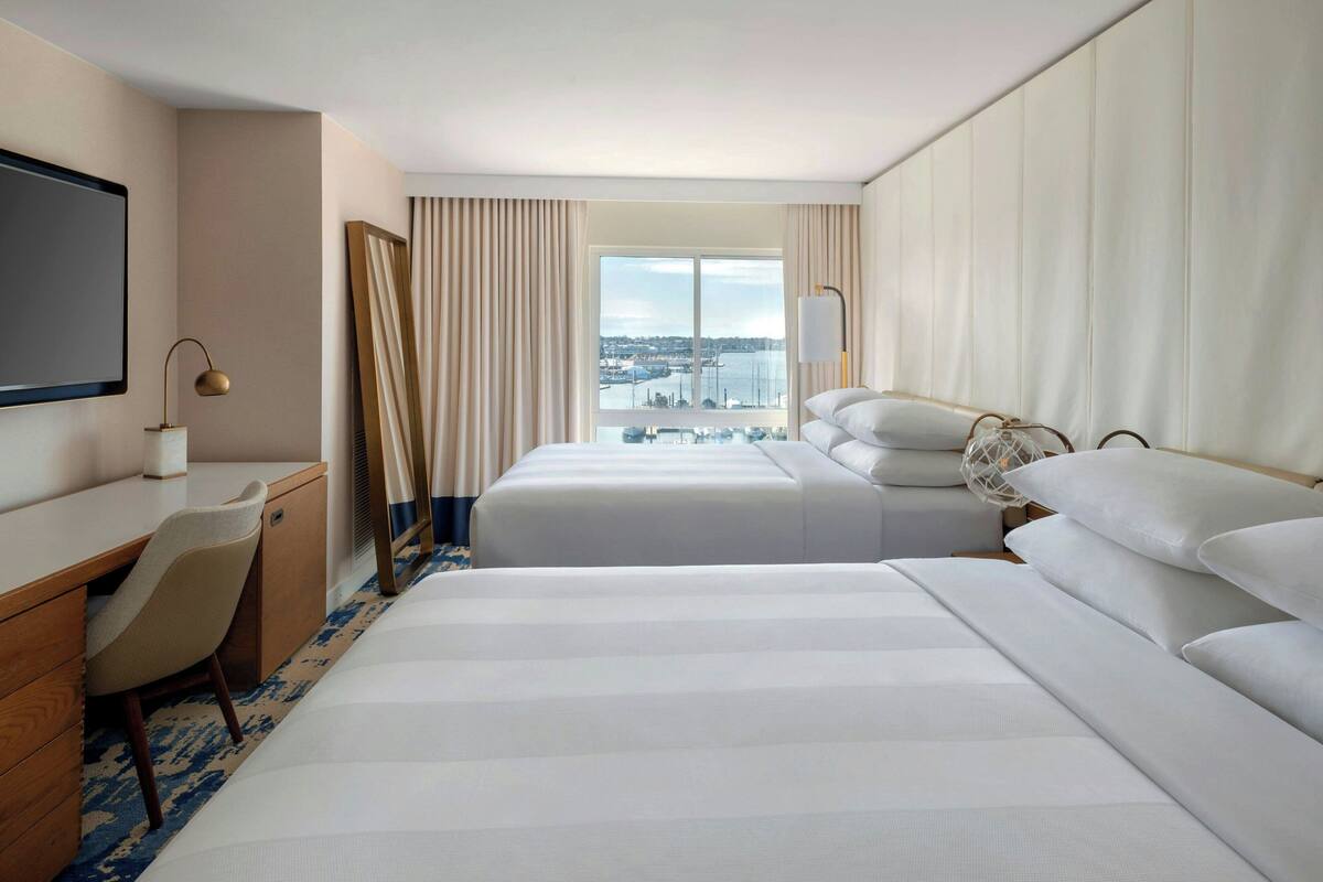 A hotel room with two beds, a desk, a chair, a TV, and a large window with a city view.