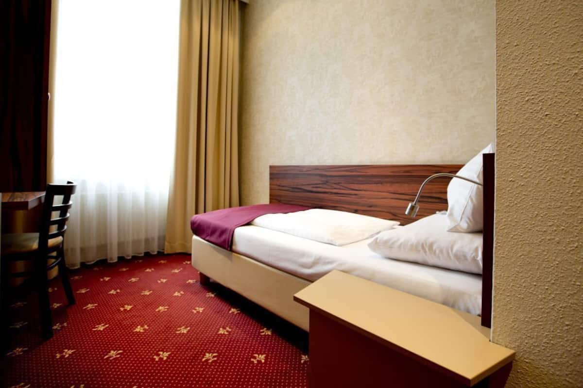 A hotel room with a bed, a wooden headboard, a desk, and a window with curtains.