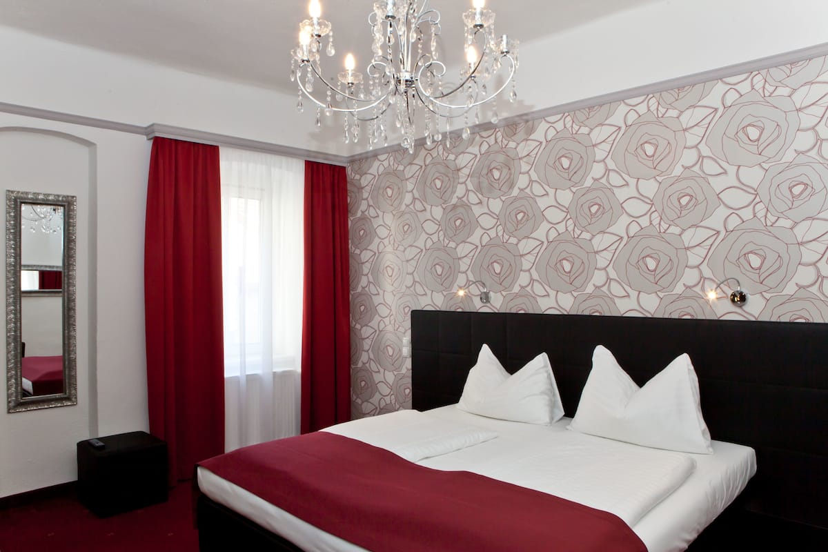 A hotel room with a large bed, red curtains, a chandelier, a mirror, and a patterned wall.
