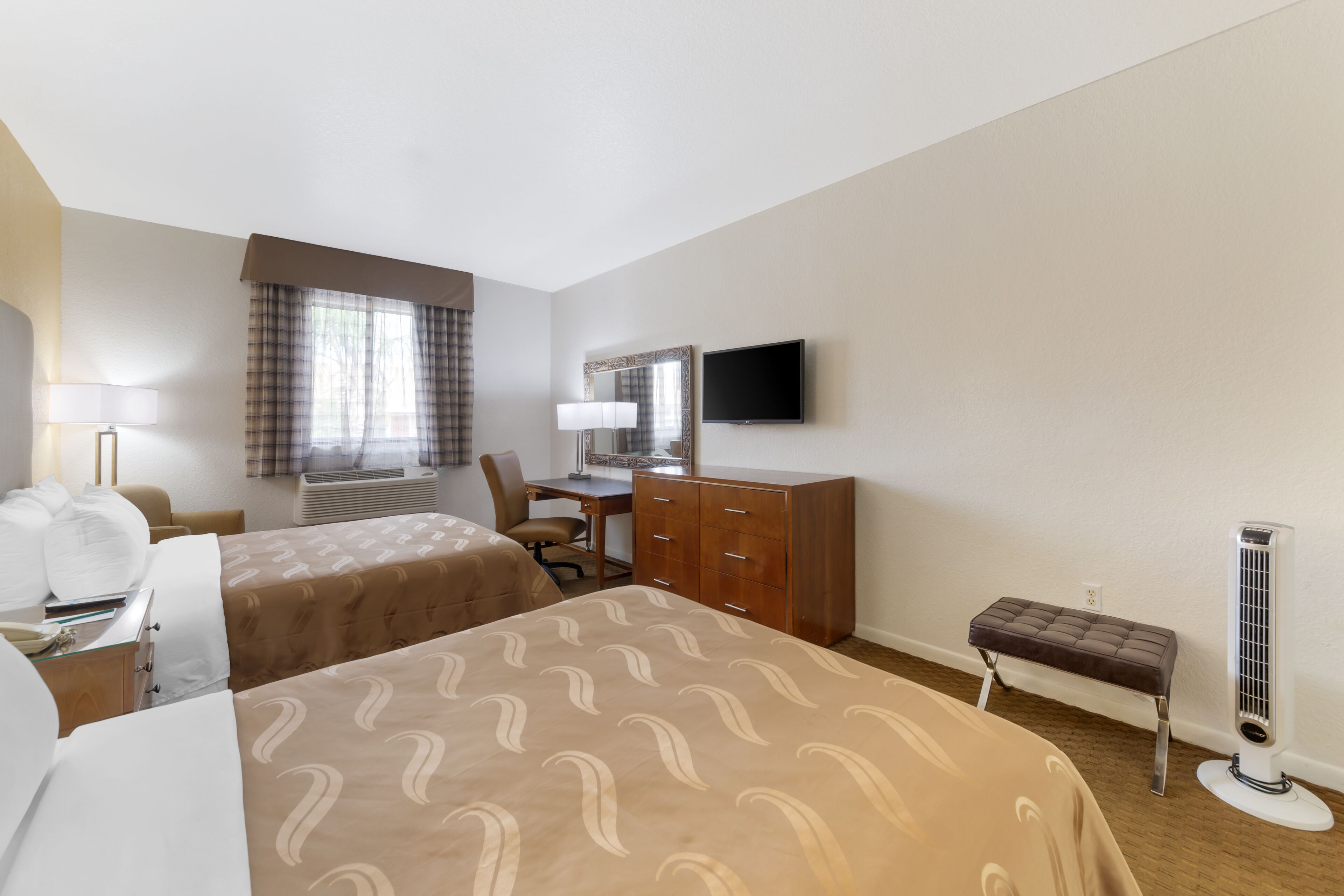A hotel room with two beds, a desk, a chair, a dresser, a television, a bench, and a standing heater.