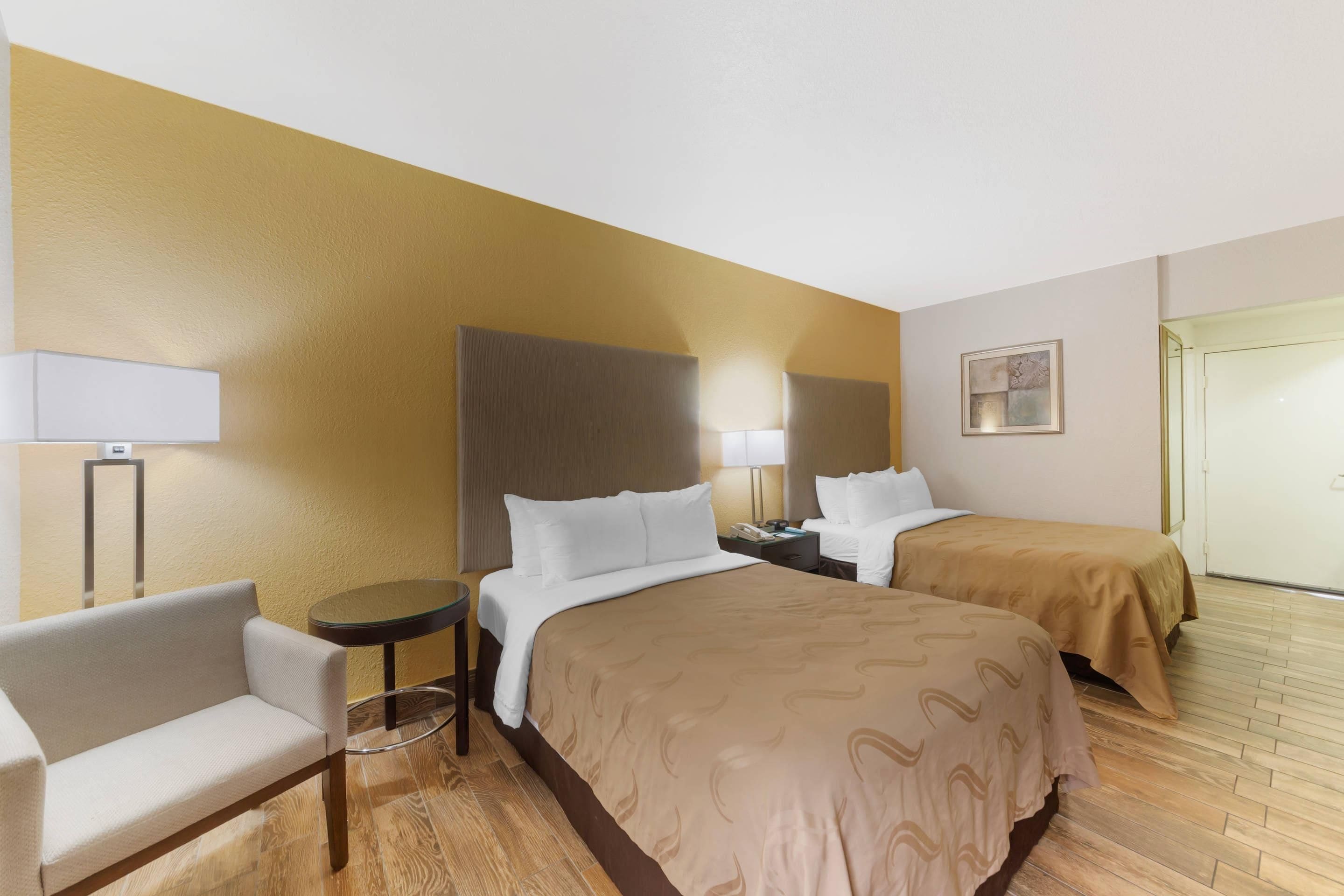 A hotel room with two beds, a small table, a chair, a lamp, and a painting on the wall.