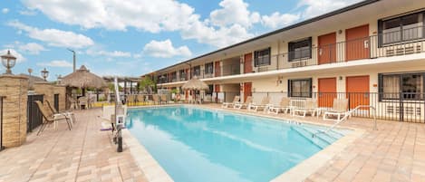 A hotel with a swimming pool, lounge chairs, and a thatched-roof structure.