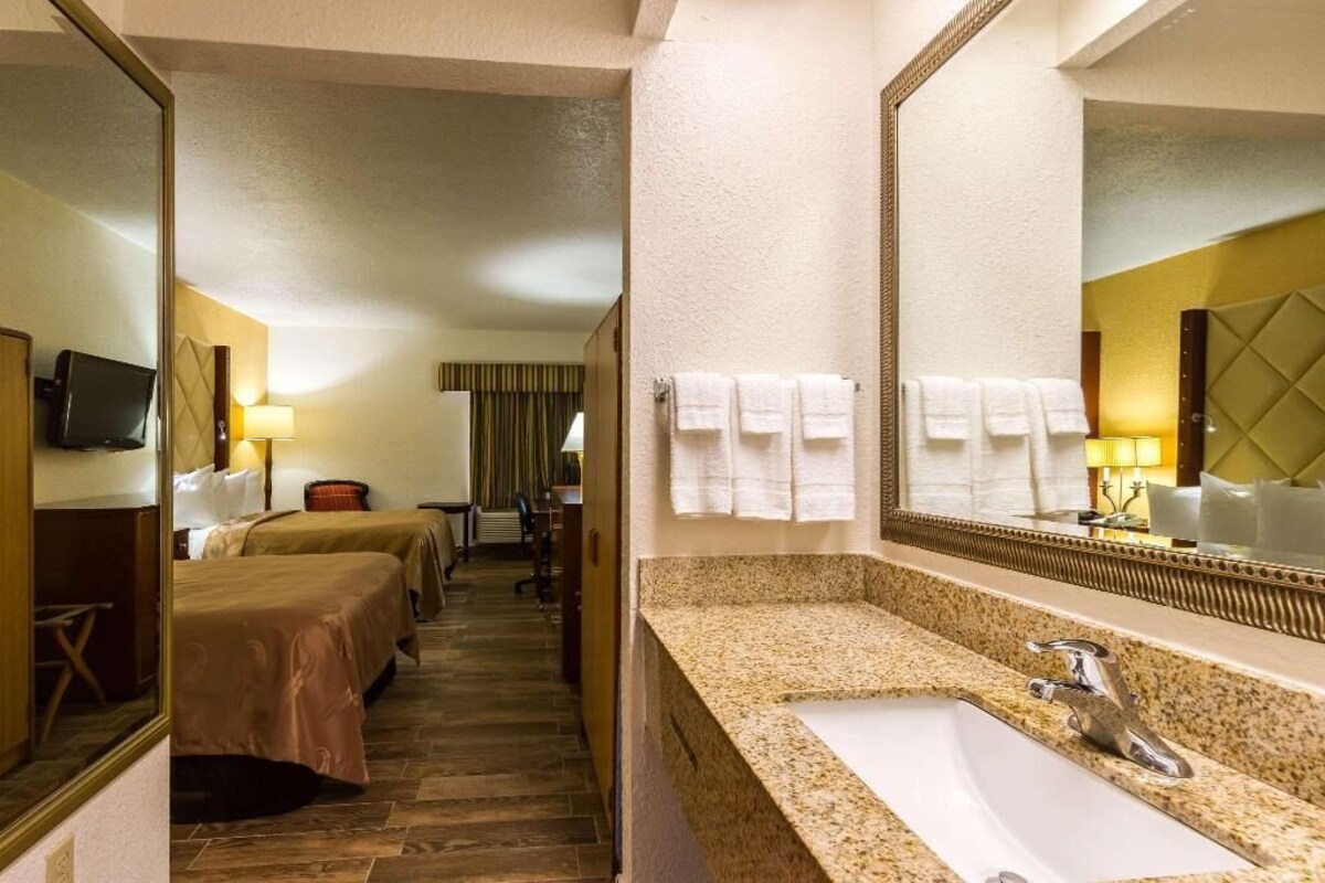 Une chambre d’hôtel avec un grand miroir, un lavabo, deux lits, une télévision et un bureau avec une lampe.