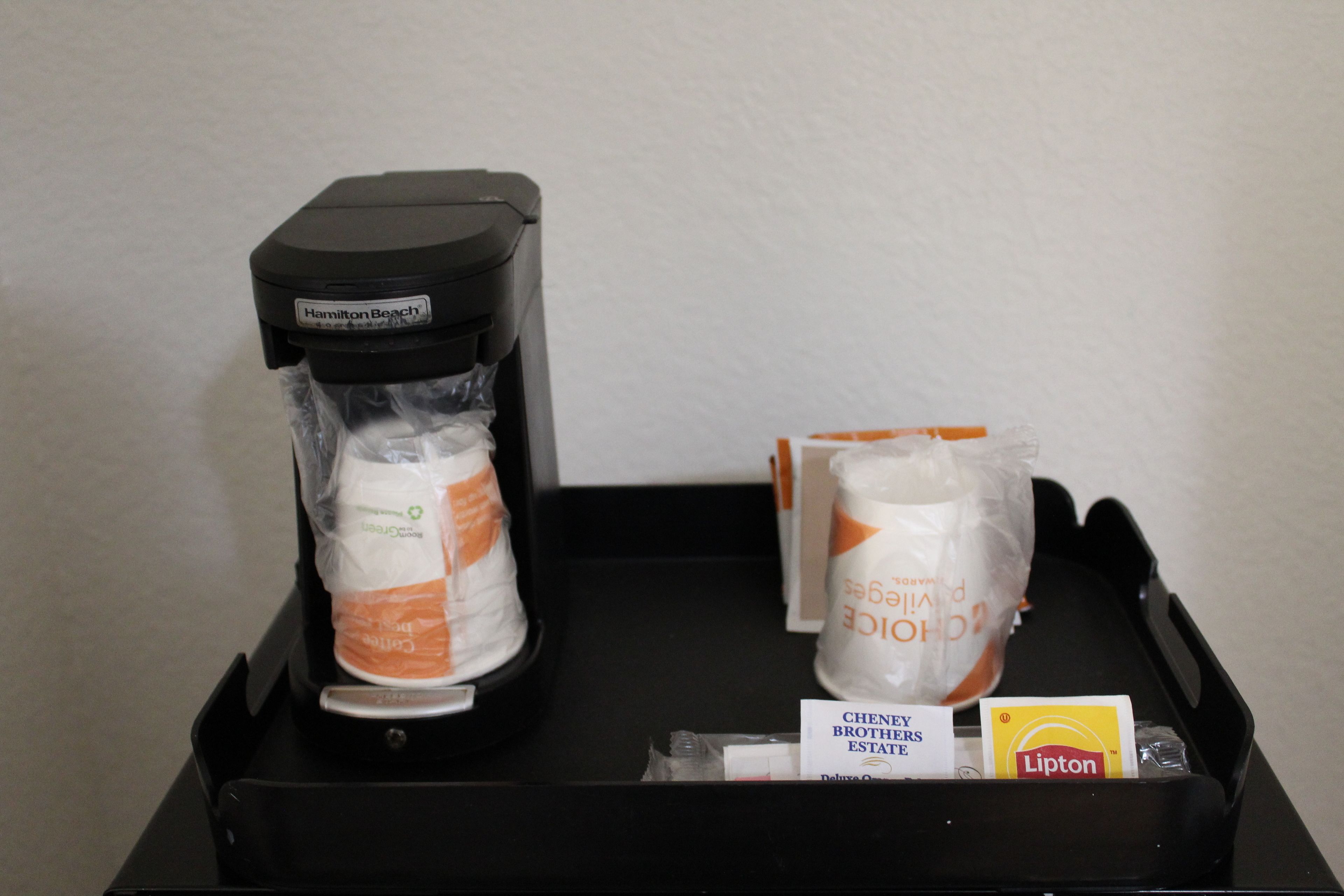 Musta tarjotinalusta, jonka päällä on Hamilton Beach -kahvinkeitin, kaksi rullaa wc-paperia, Lipton-teepakkaus ja Cheney Brothers Estate -kyltti.