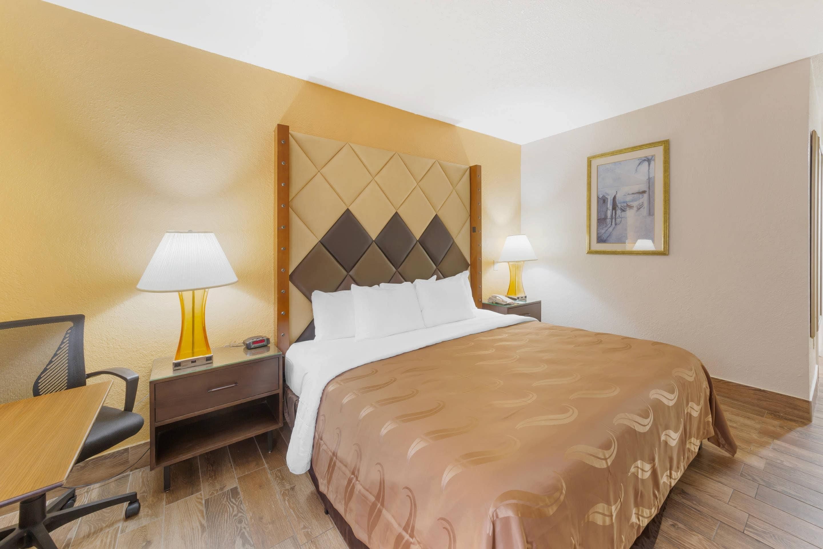 Una habitación de hotel con una cama, dos mesitas de noche con lámparas, una silla, un escritorio y un cuadro en la pared.