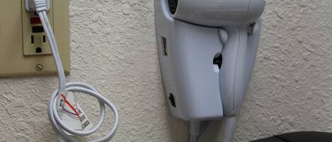Un assecador de cabells fixat a la paret, amb el cable d'alimentació connectat a una presa elèctrica propera.