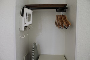 Esquina de una habitación con una pared blanca, un estante de madera que sostiene un electrodoméstico blanco y un juego de herramientas con mangos de madera, y una silla a rayas.