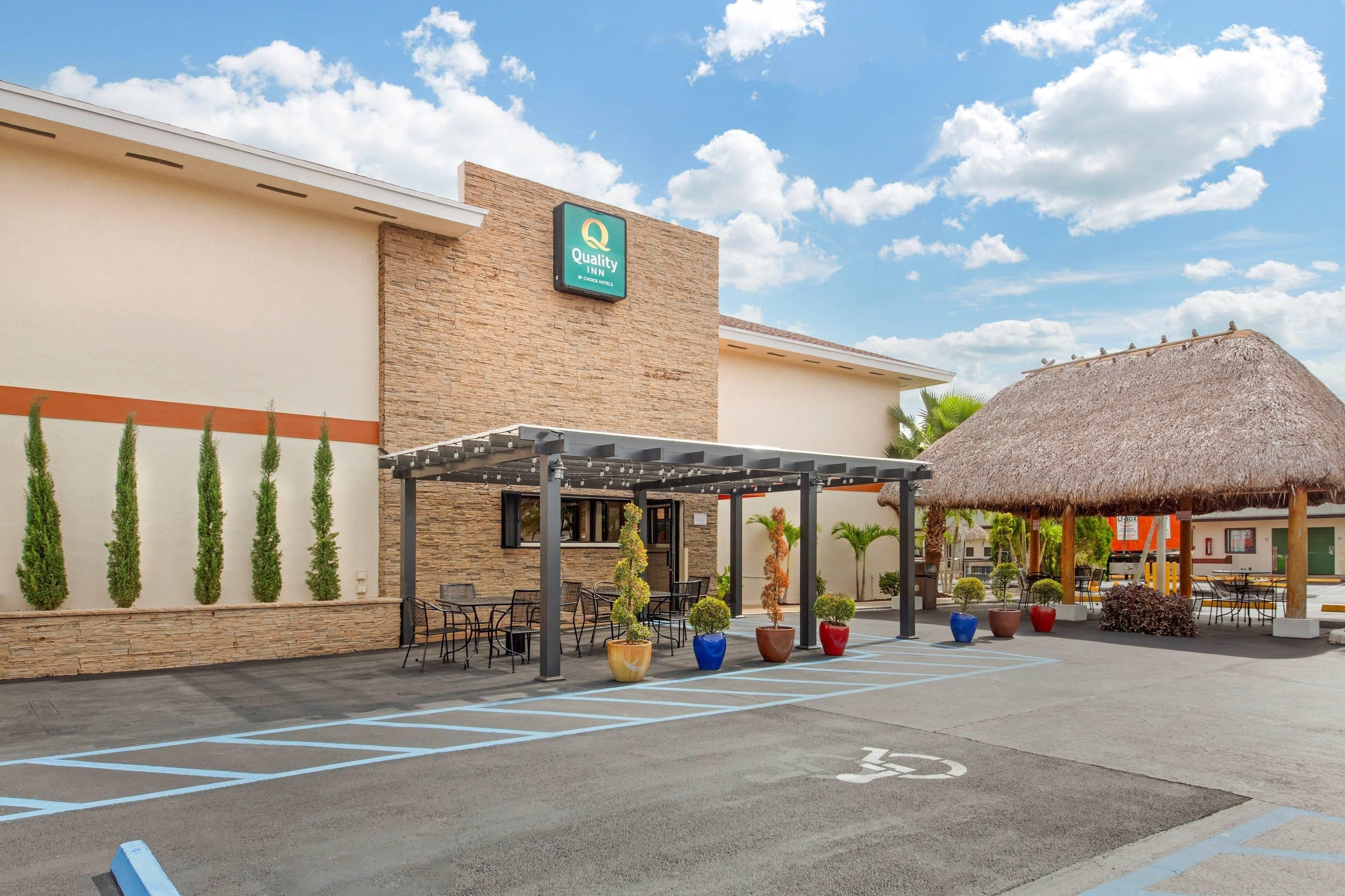 A hotel entrance with a covered outdoor seating area, a thatched-roof structure, and a parking lot with marked spaces.
