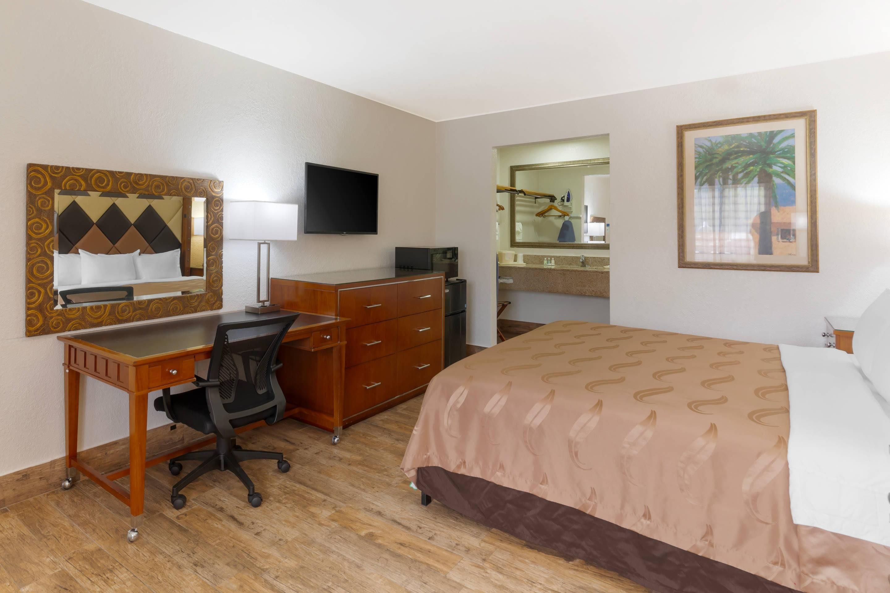 A hotel room with a bed, desk, chair, dresser, television, and a bathroom with a mirror and sink.