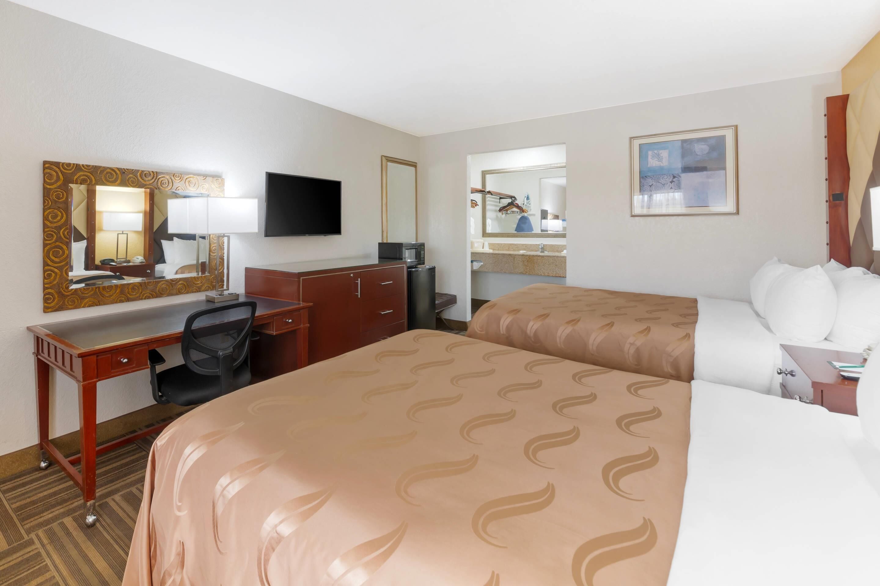 A hotel room with a bed, a desk, a chair, a television, a mirror, and a bathroom with a sink and mirror.