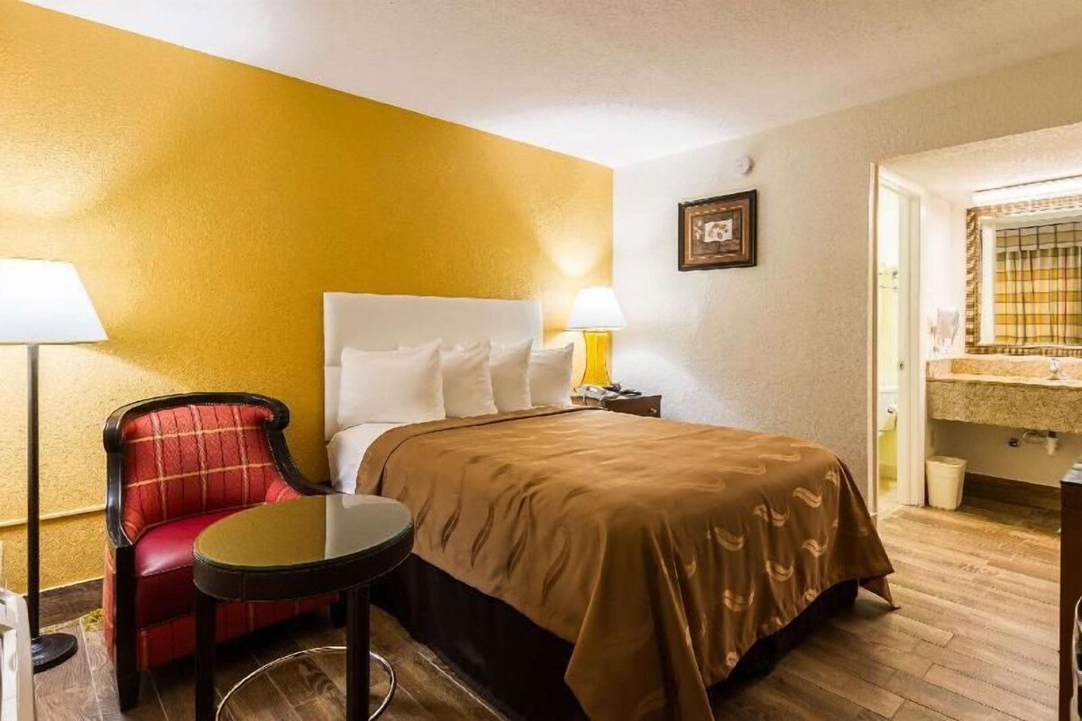 A hotel room with a bed, a red chair, a small round table, a nightstand, and a bathroom with a mirror and sink.