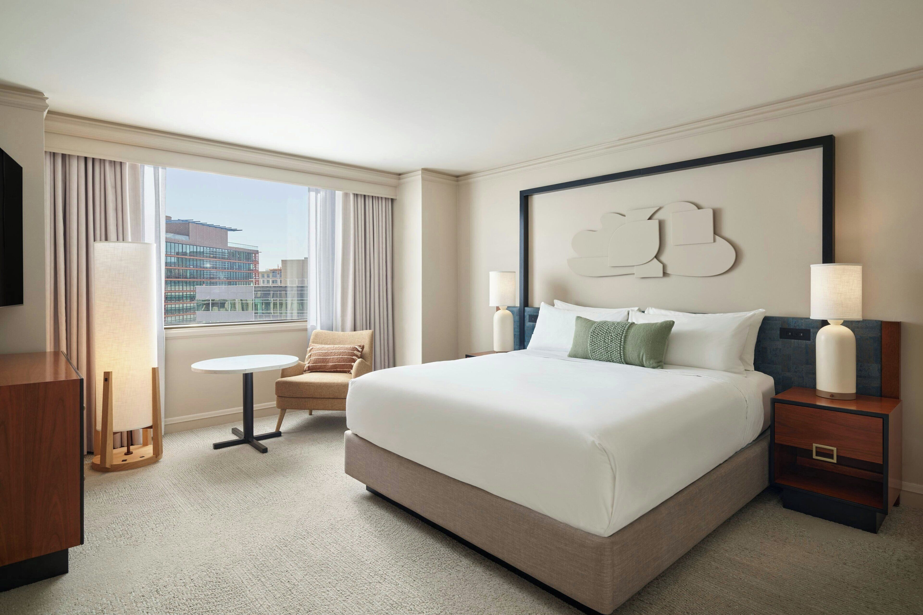 A hotel room with a large bed, a small round table, a chair, and a view of the cityscape through the window.