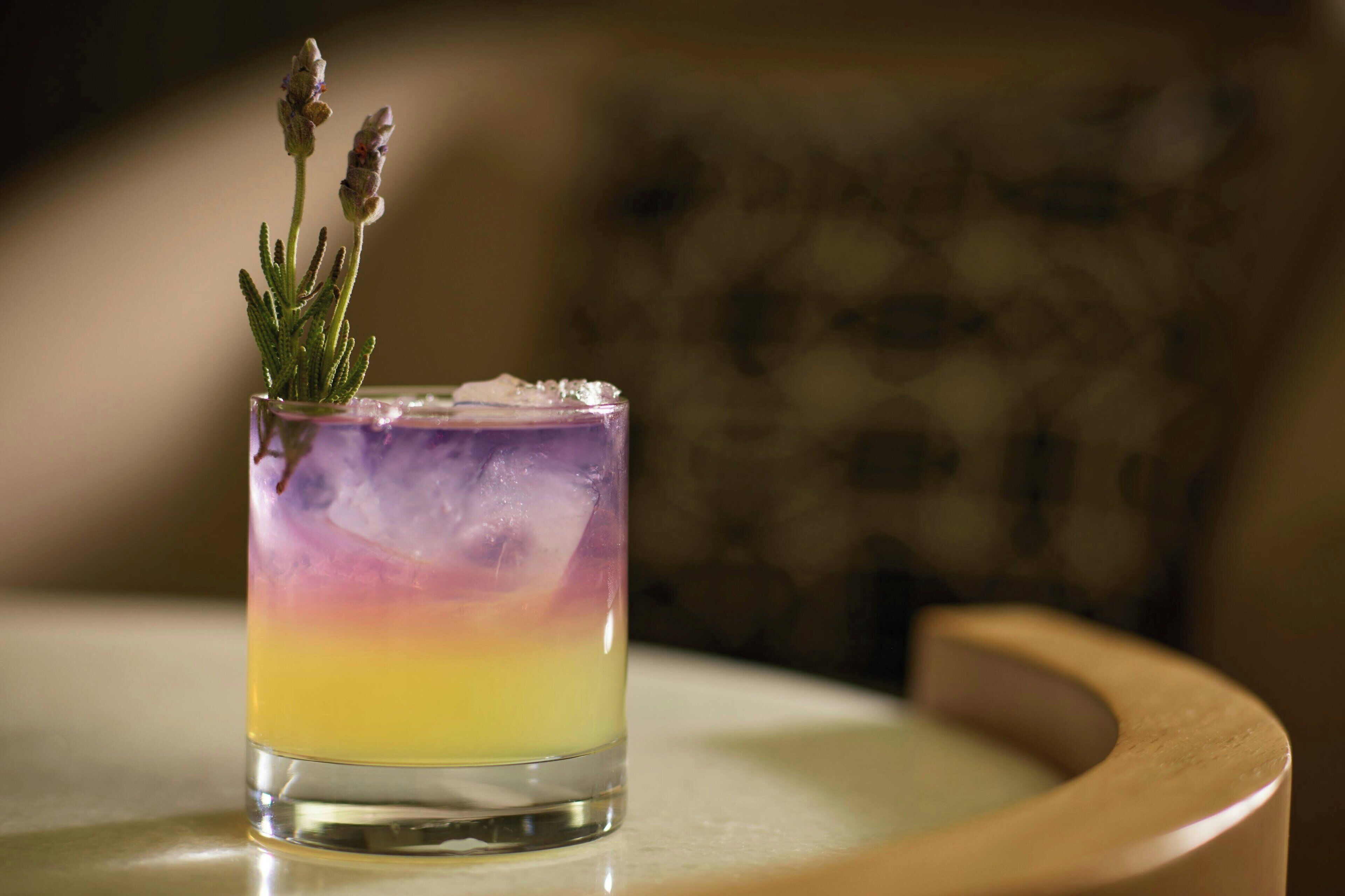 A cocktail with a gradient color effect, ice cubes, and a sprig of rosemary on a glass.