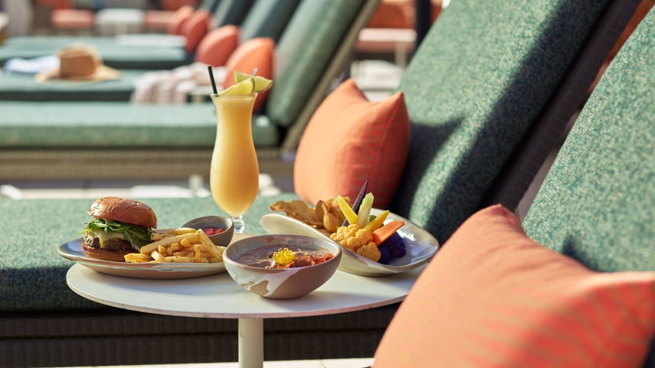 A dining table with a burger, fries, a drink, and a bowl of soup in a poolside setting.