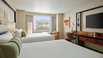 A hotel room with two beds, a desk, a chair, a TV, and a window with a city and mountain view.