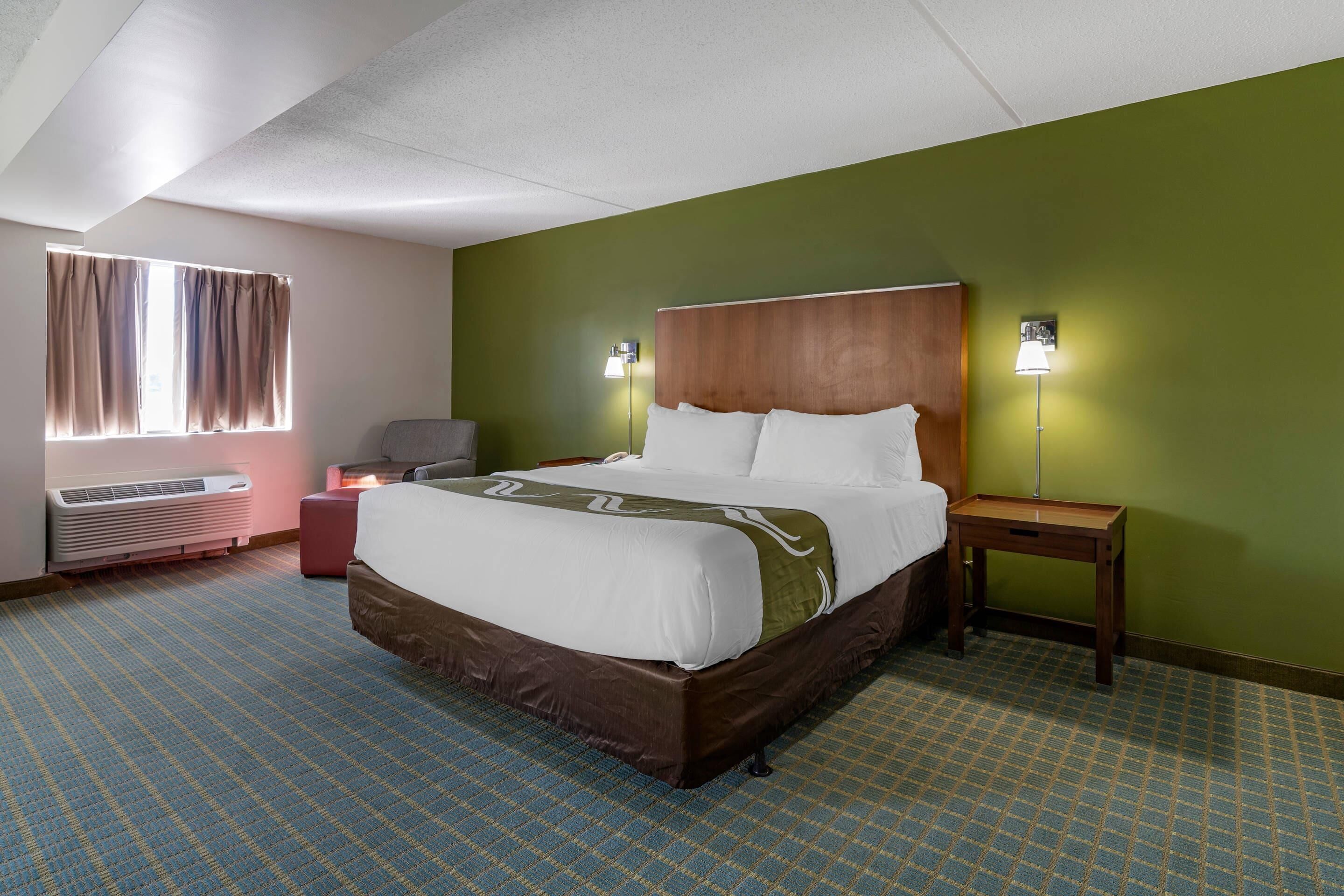 A hotel room with a large bed, a bedside table, a lamp, a chair, and a window with curtains.