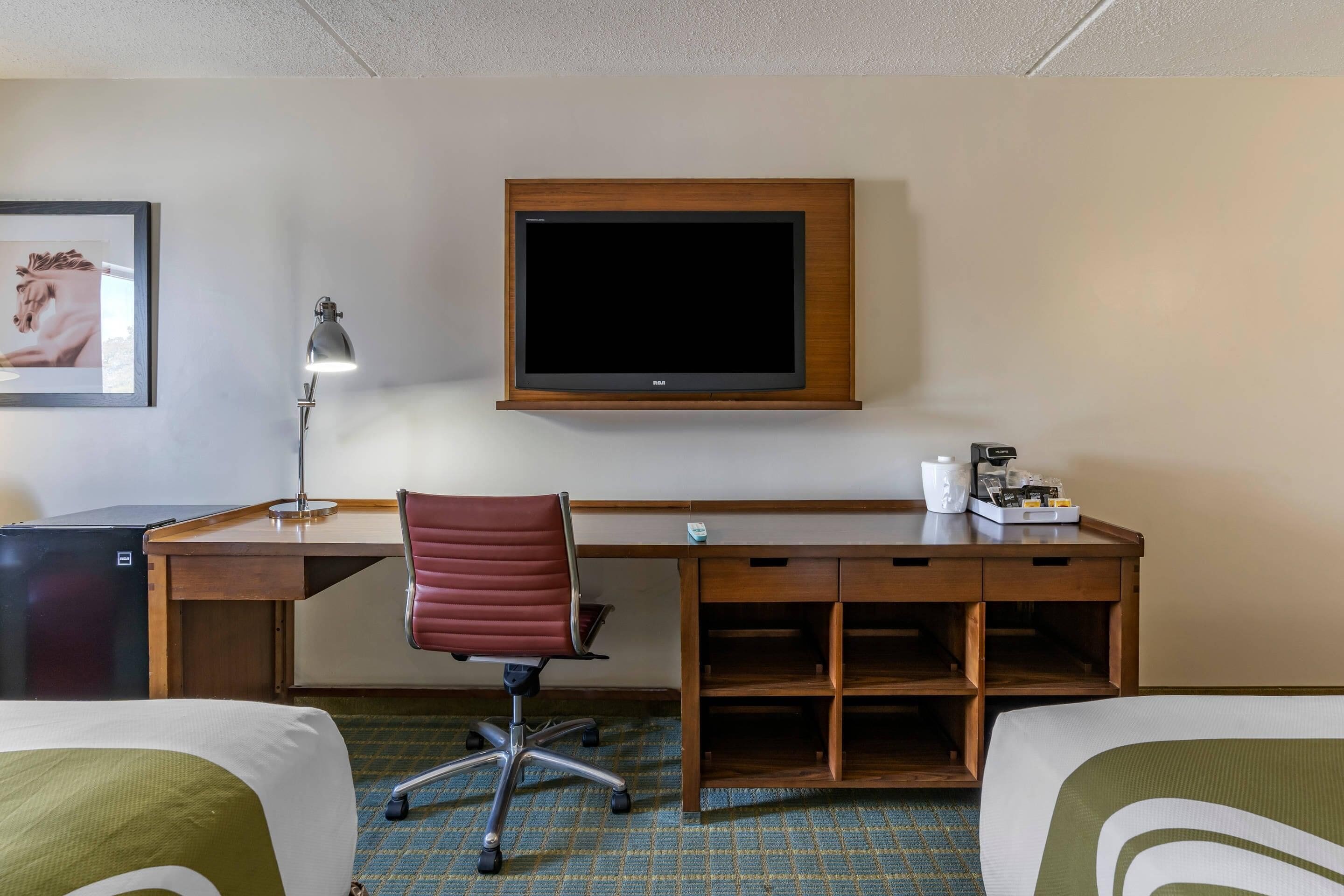 A hotel room with a bed, desk, chair, TV, and a small table with a cup and tissues.