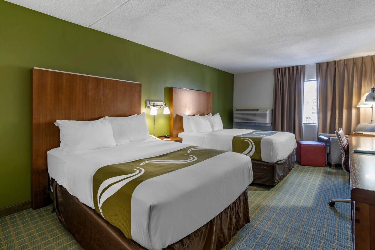 A hotel room with two beds, a desk, a chair, a lamp, and a window with curtains.