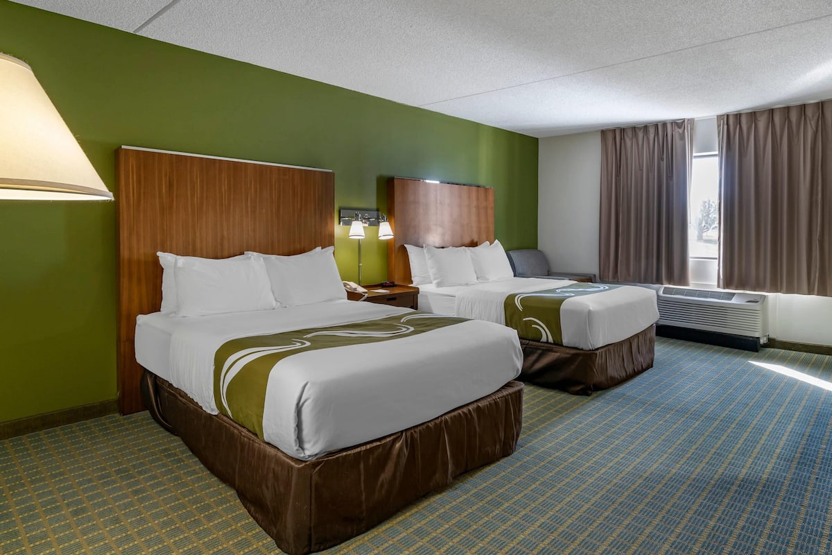 A hotel room with two beds, a green wall, a patterned carpet, and a window with curtains.