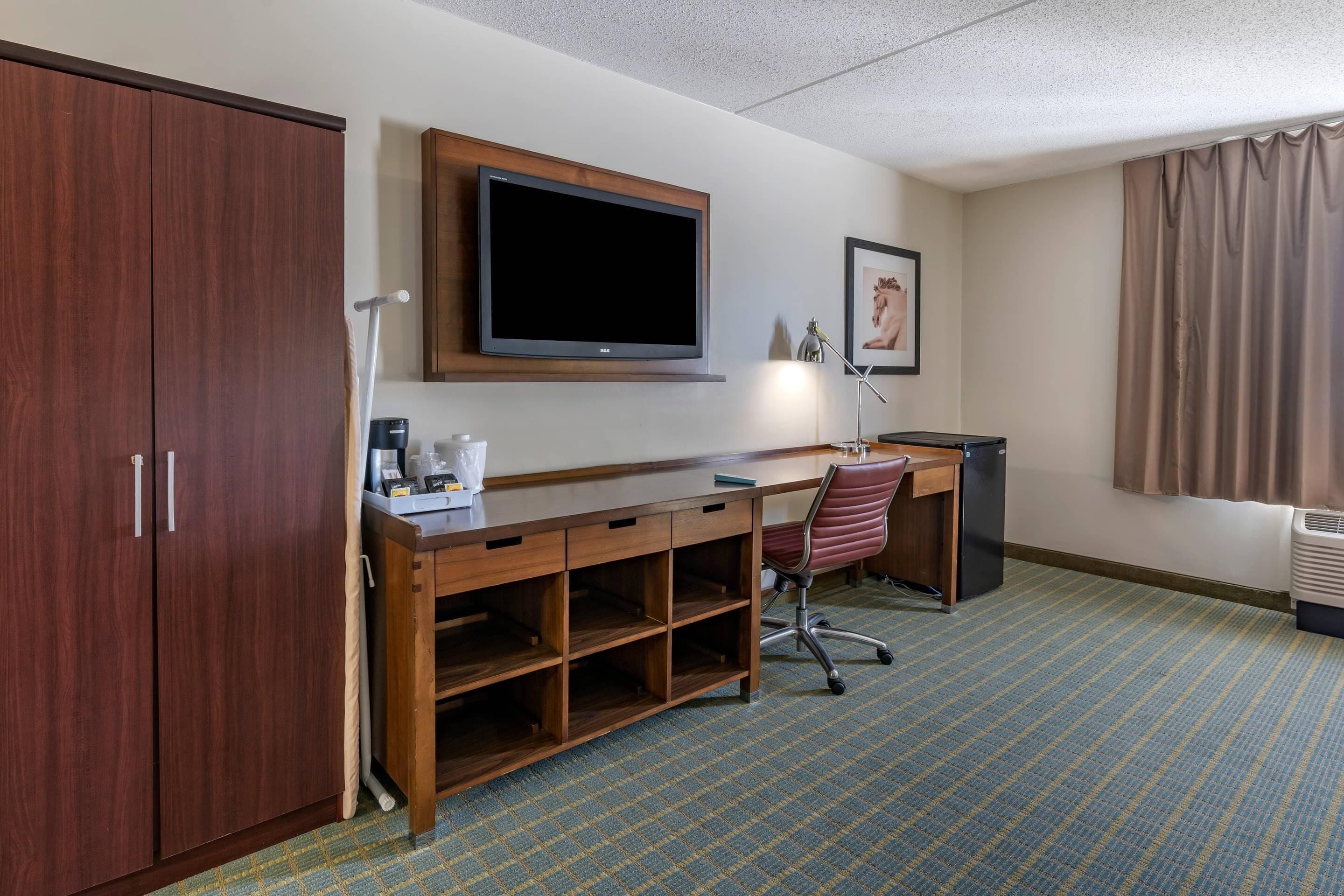 A hotel room with a wooden desk, a television mounted above it, a chair, a wardrobe, and a patterned carpet.