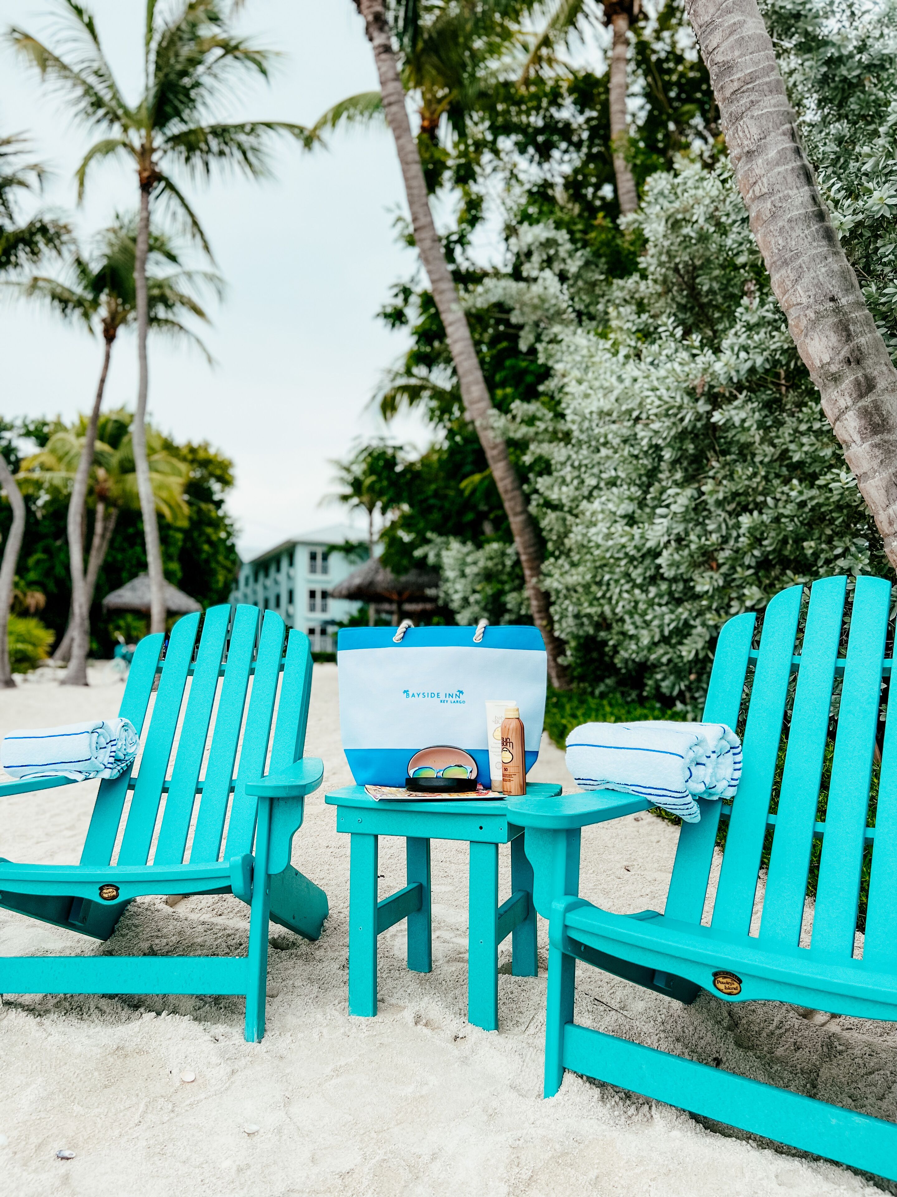 Two turquoise beach chairs with towels, a bag, and a bottle on a small table on a sandy beach with palm trees in the background.