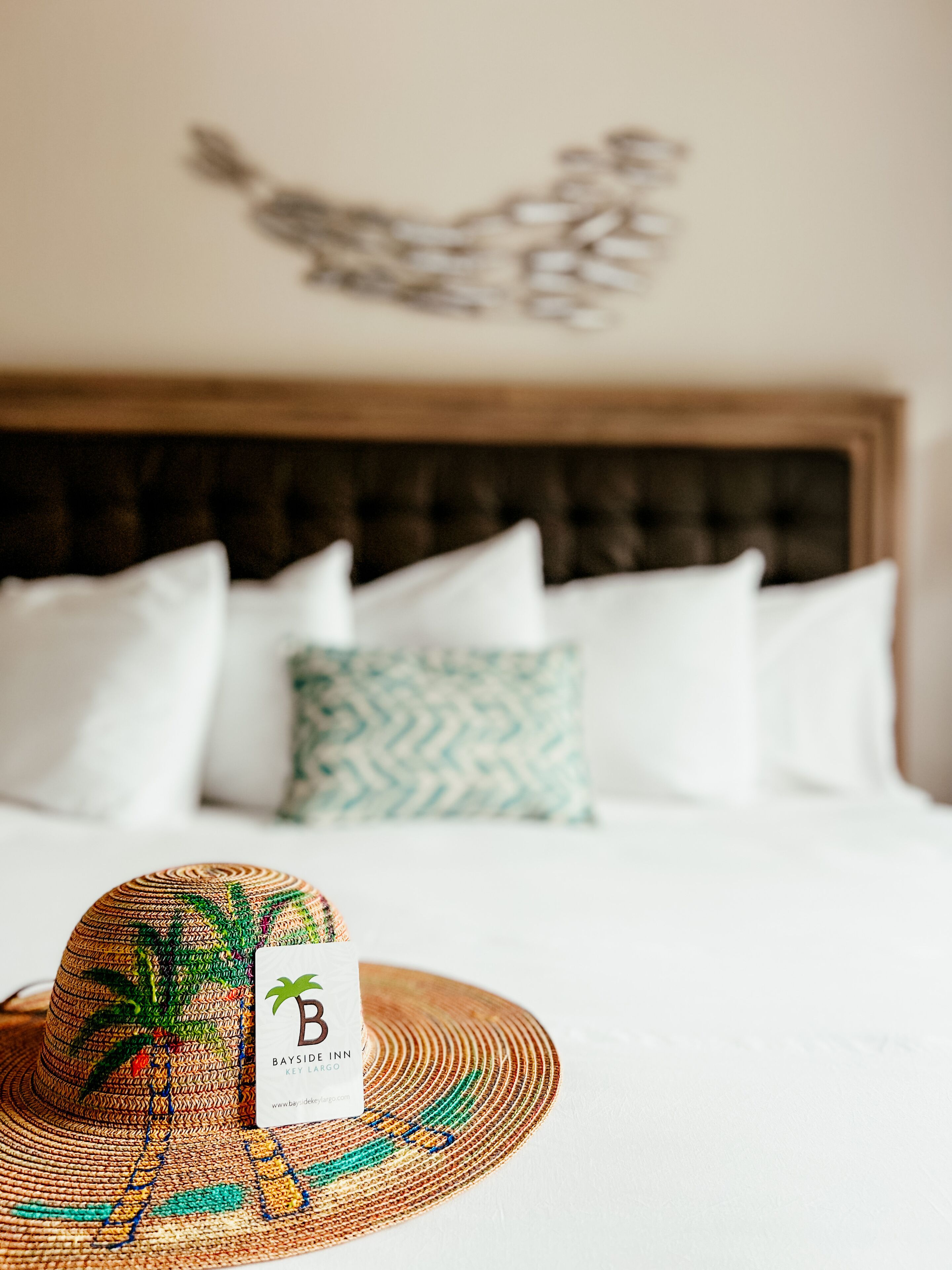 Una cama con almohadas blancas y una almohada verde estampada, un sombrero tejido con un logotipo y una obra de arte en la pared sobre la cama.