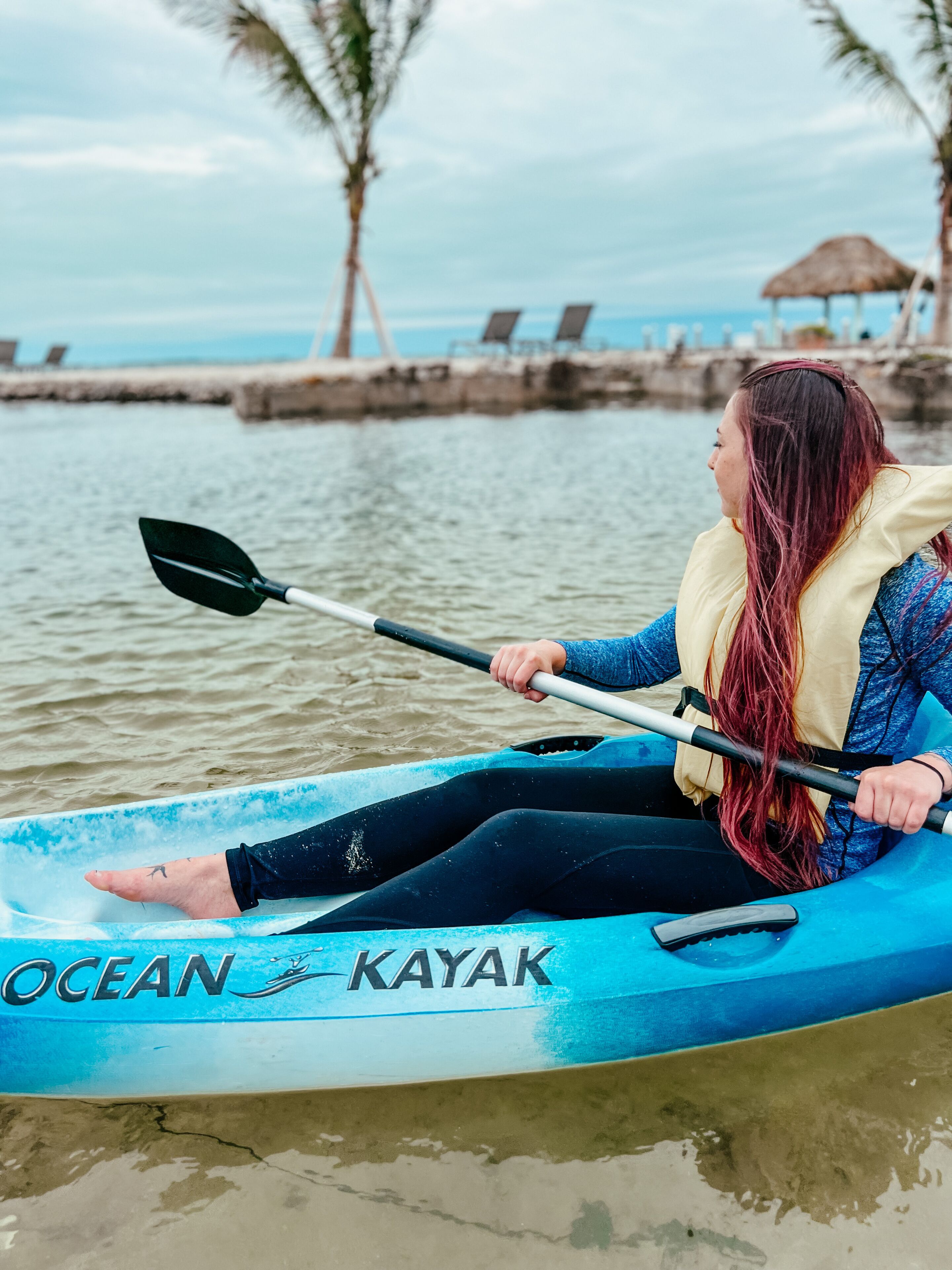 A person kayaking on a calm body of water with a thatched-roof structure in the background.