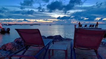 Una playa con dos sillas de descanso y una mesa, un muelle con barcos, y un atardecer sobre el agua.