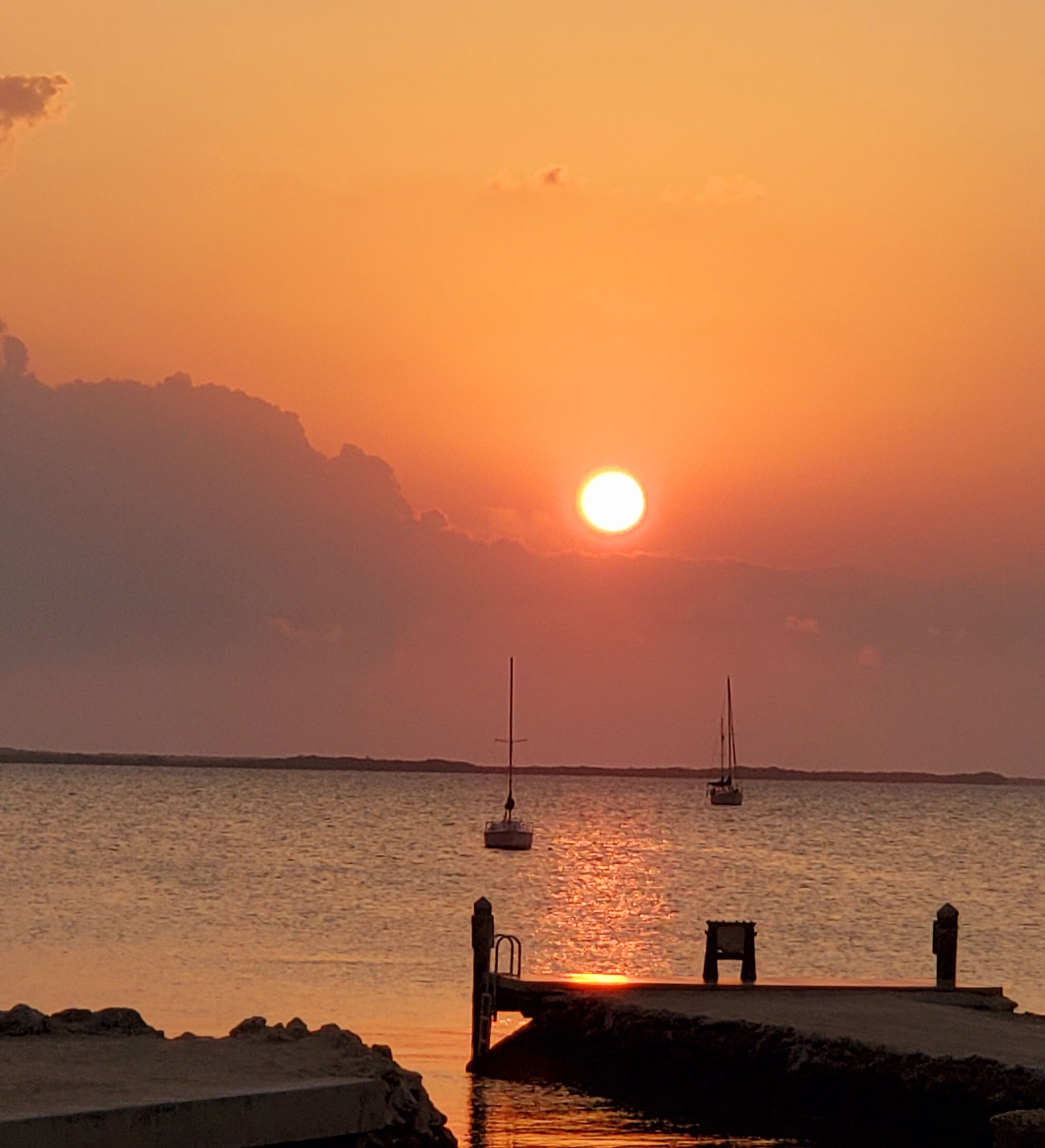 A sunset over a calm sea with two sailboats anchored. A pier extends into the water with a small dock and a chair.
